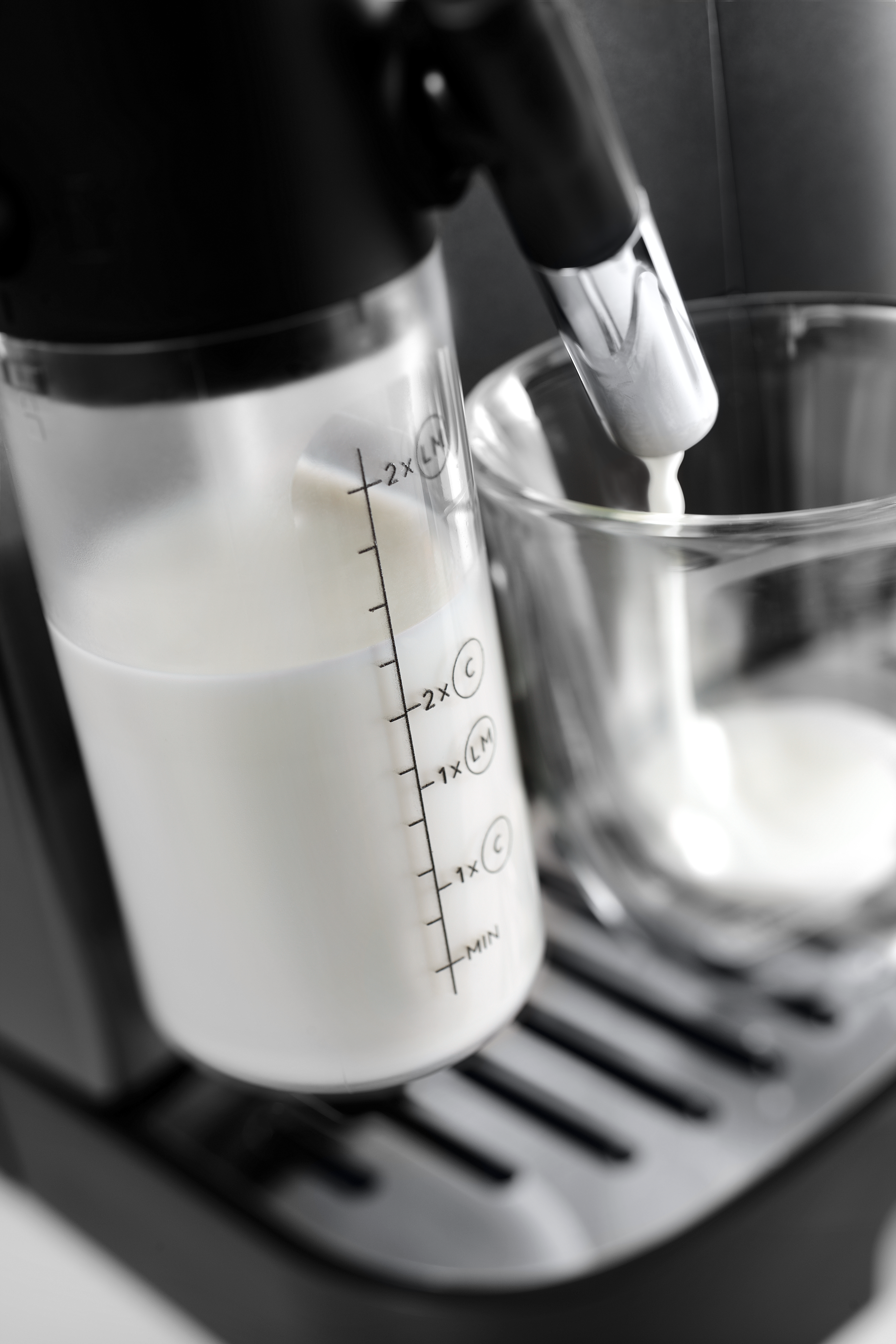 Milch wird aus einem Milchaufschäumer in ein Glas gegossen, Kaffeemaschine im Bild.