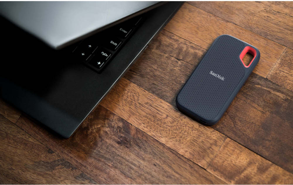 Un ordinateur portable noir est partiellement ouvert. Un disque dur externe noir et rouge repose sur du bois.