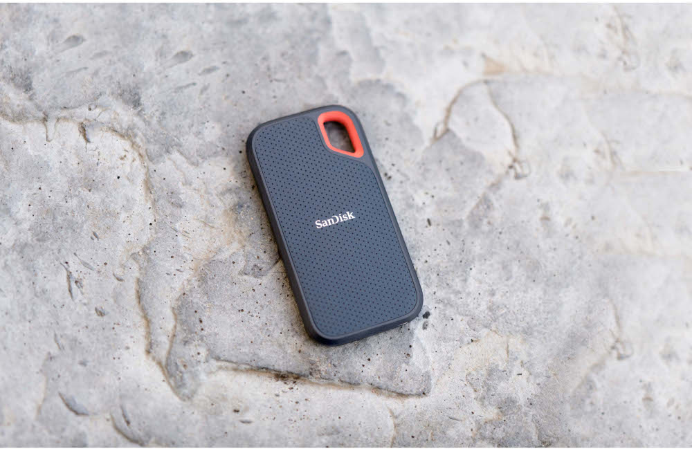 SSD portable SanDisk gris foncé sur une surface en pierre rugueuse. Bord rouge en haut.