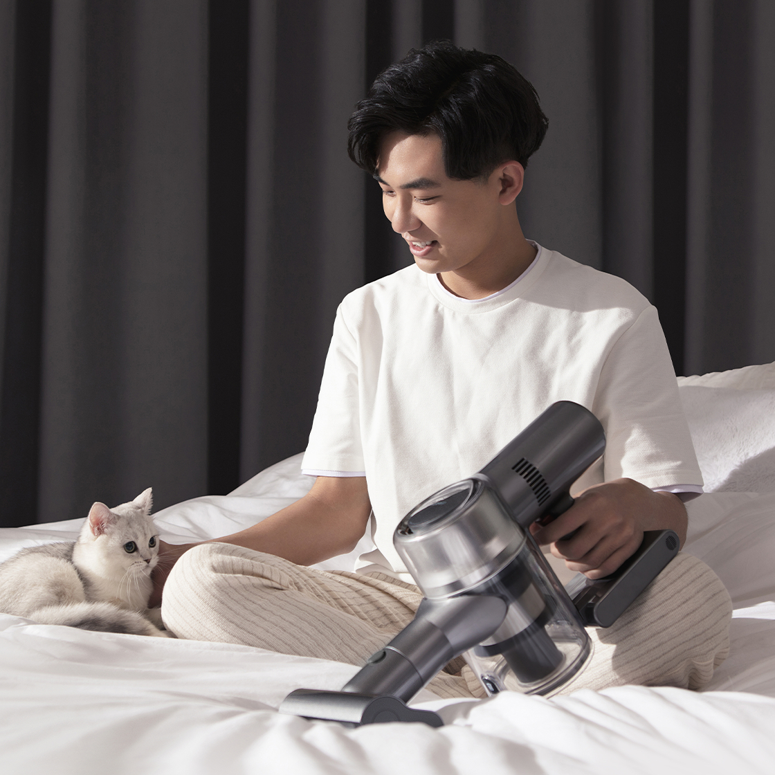 Ein Mann sitzt auf einem Bett, streichelt eine Katze und hält einen grauen Staubsauger.