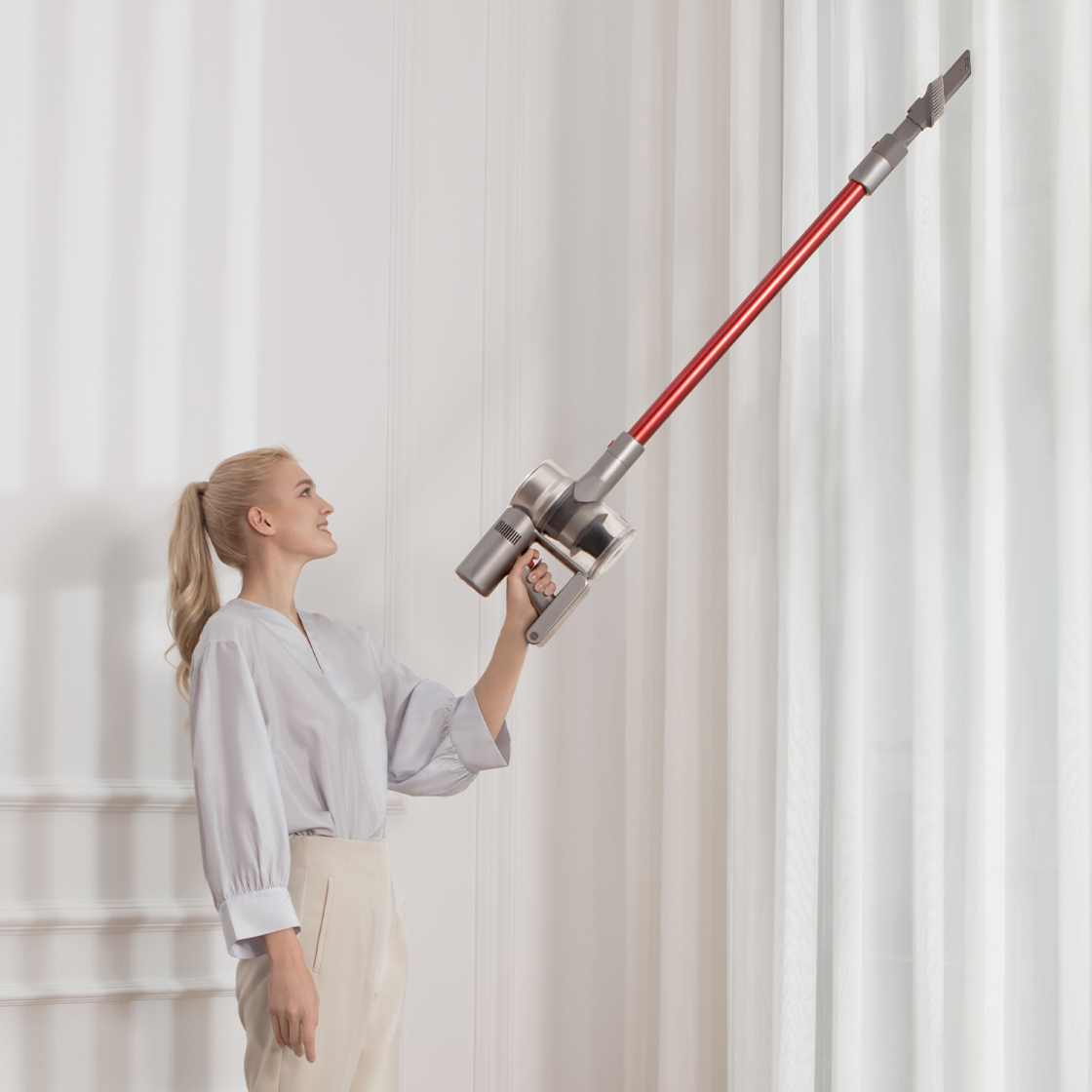 Eine Frau hält einen rot-silbernen Staubsauger, um eine Wand zu reinigen. Sie lächelt.