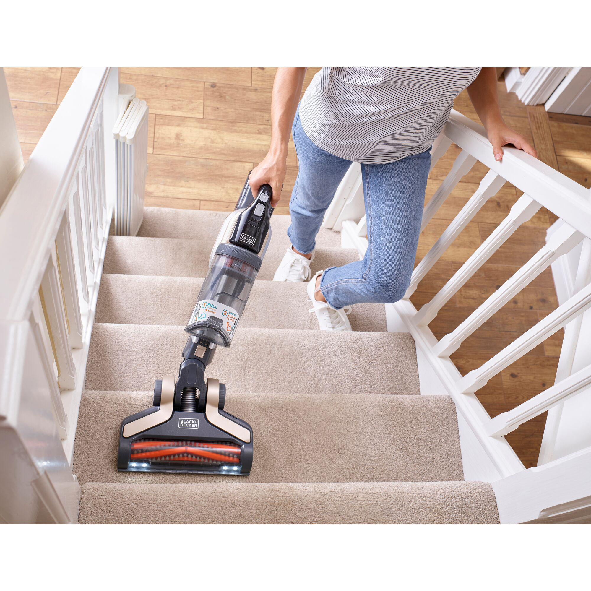 Frau, die Treppen mit einem BLACK+DECKER-Staubsauger saugt. Beiger Teppich, weißes Geländer.