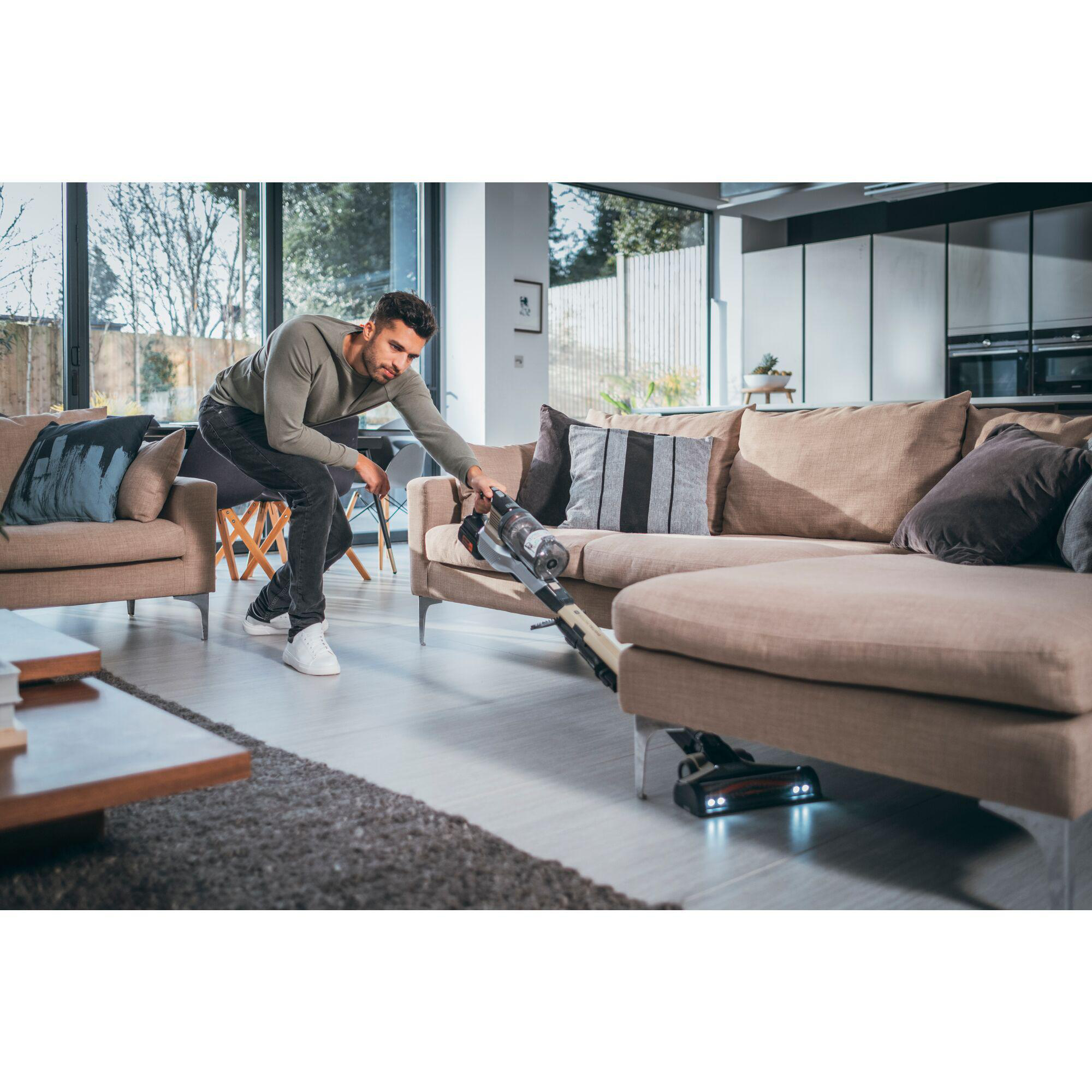 Mann, der mit einem Handstaubsauger einen Sofa saugt. Der Raum hat ein Fenster und braune Möbel.
