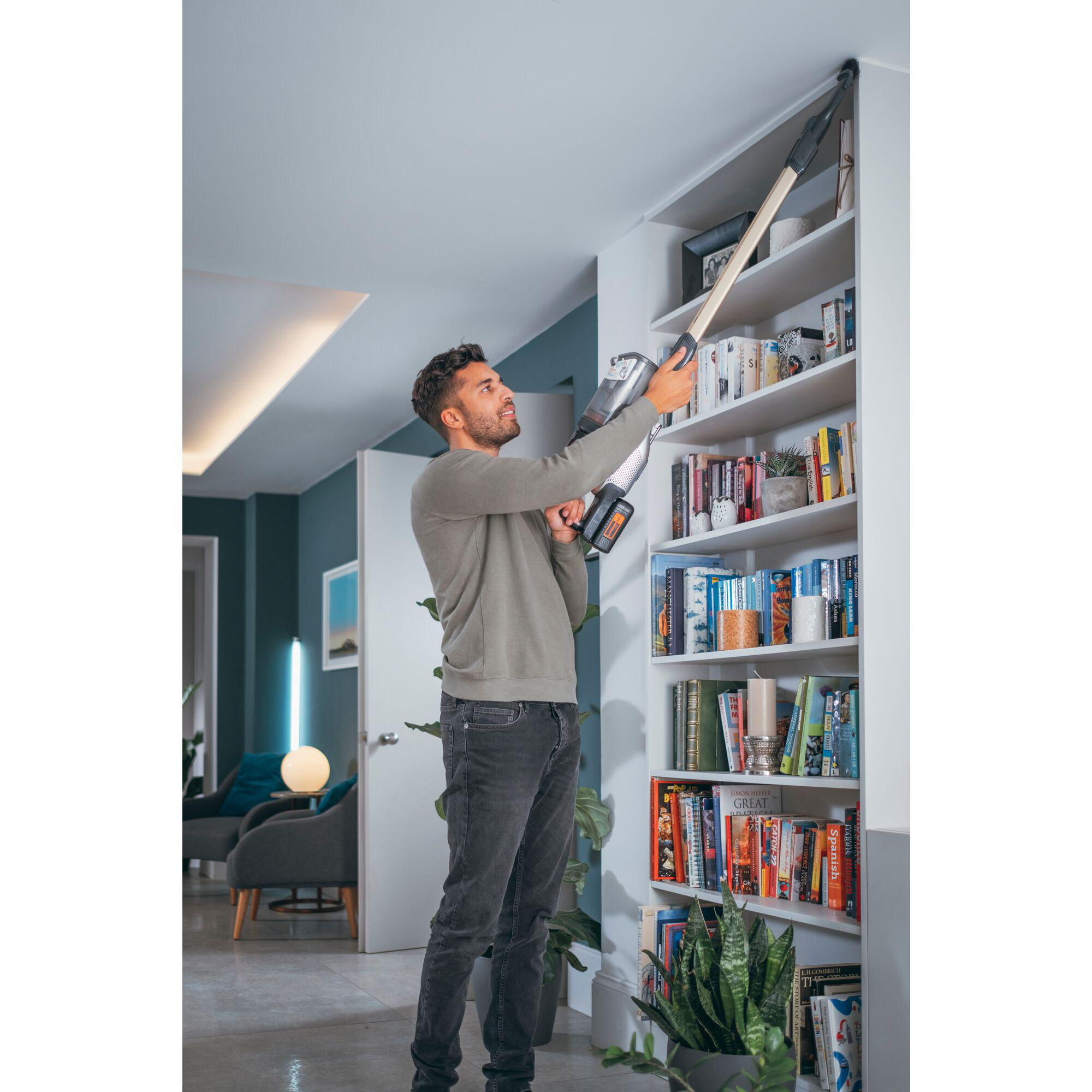 Mann, der mit einem kabellosen Staubsauger ein Bücherregal absaugt. Bücher und Dekorationen auf Regalen.