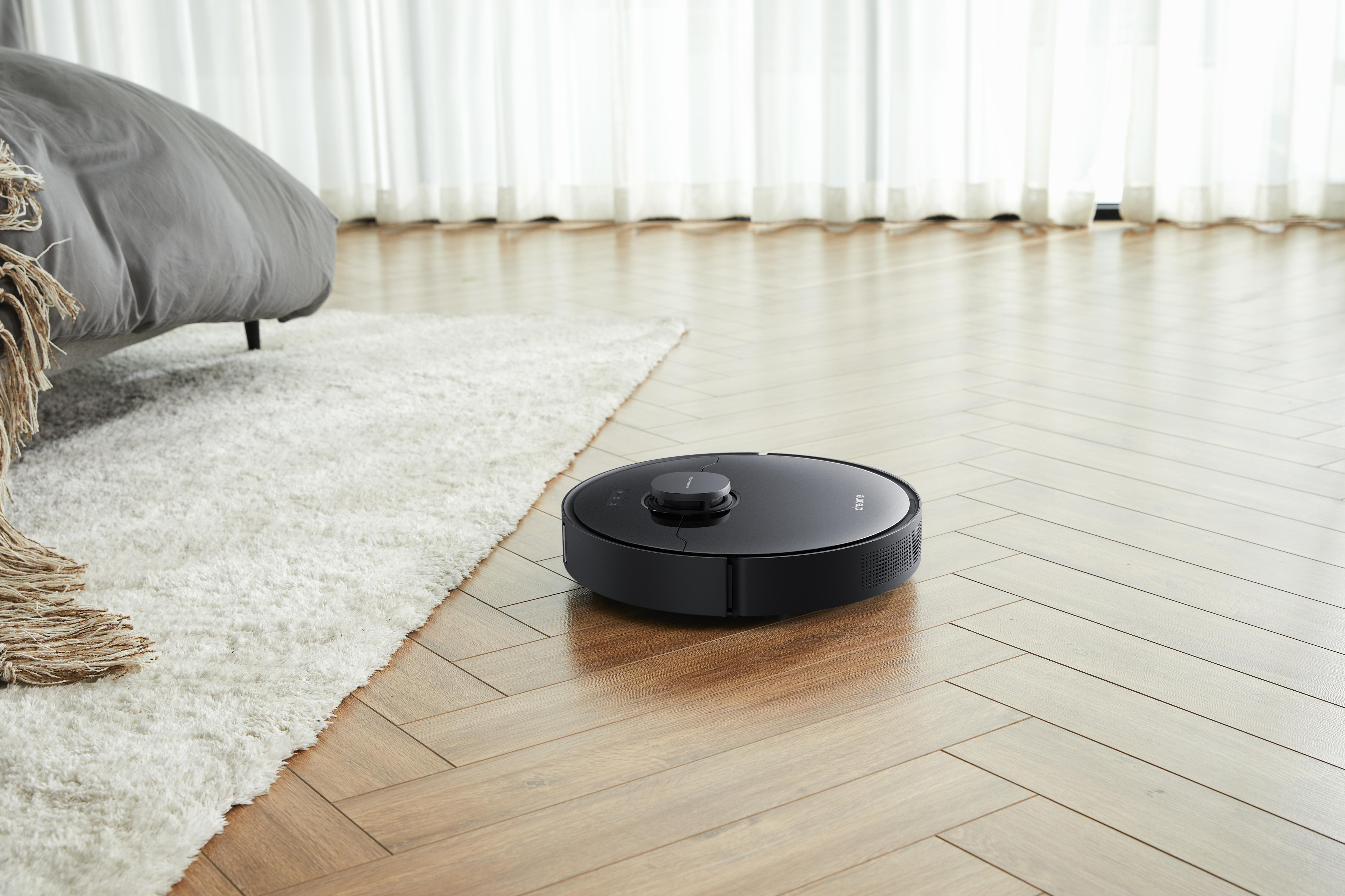 Schwarzer Roboter-Staubsauger auf Holzboden, in der Nähe eines weißen Teppichs und Bettes.