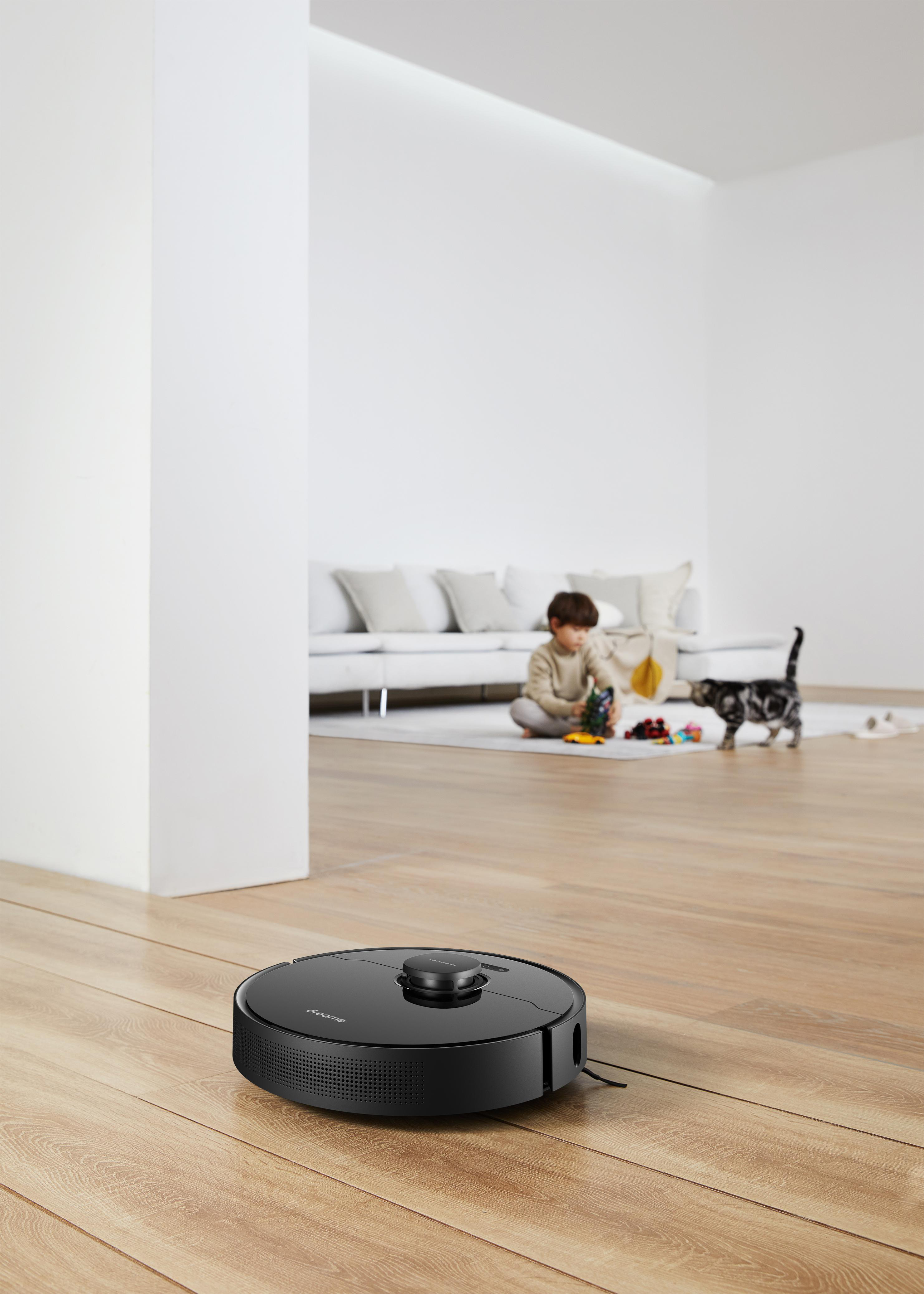 Ein schwarzer Roboter-Staubsauger auf einem Holzboden. Ein Junge und eine Katze sind im Hintergrund zu sehen.