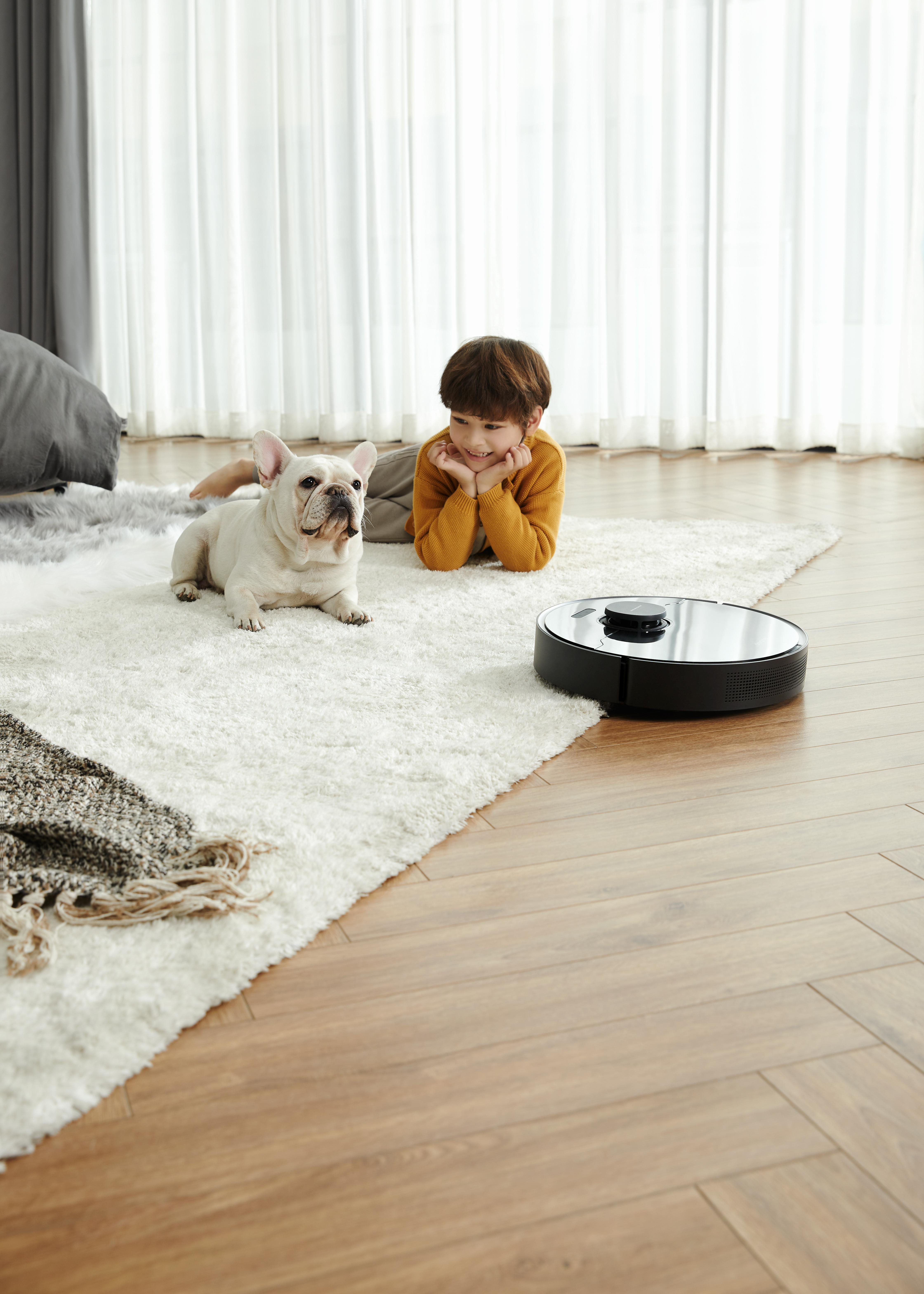 Ein Junge und ein Hund auf einem Teppich, in der Nähe befindet sich ein schwarzer Roboterstaubsauger.