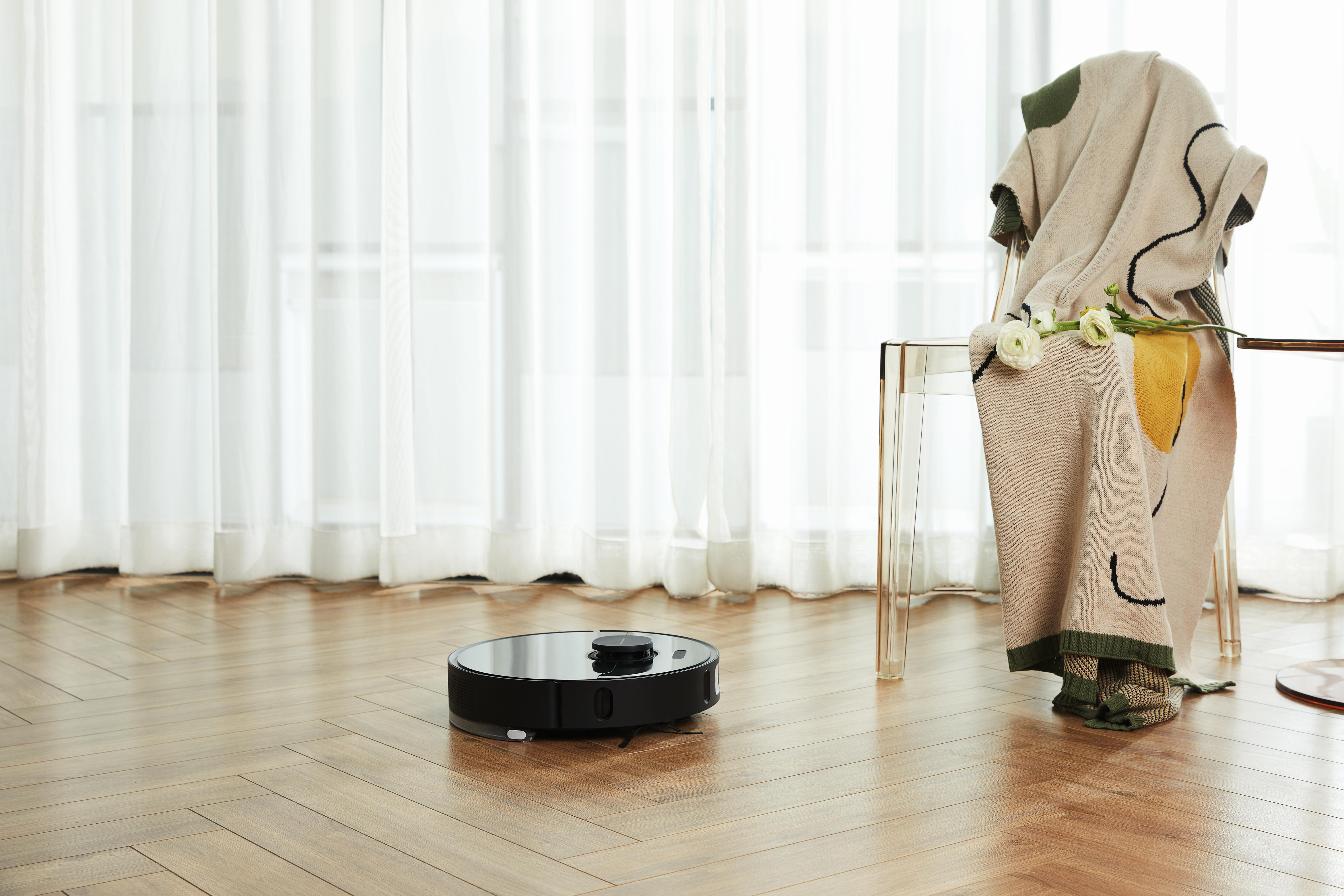 Ein schwarzer Roboter-Staubsauger auf einem Hartholzboden, mit einem Stuhl und einer Decke im Hintergrund.