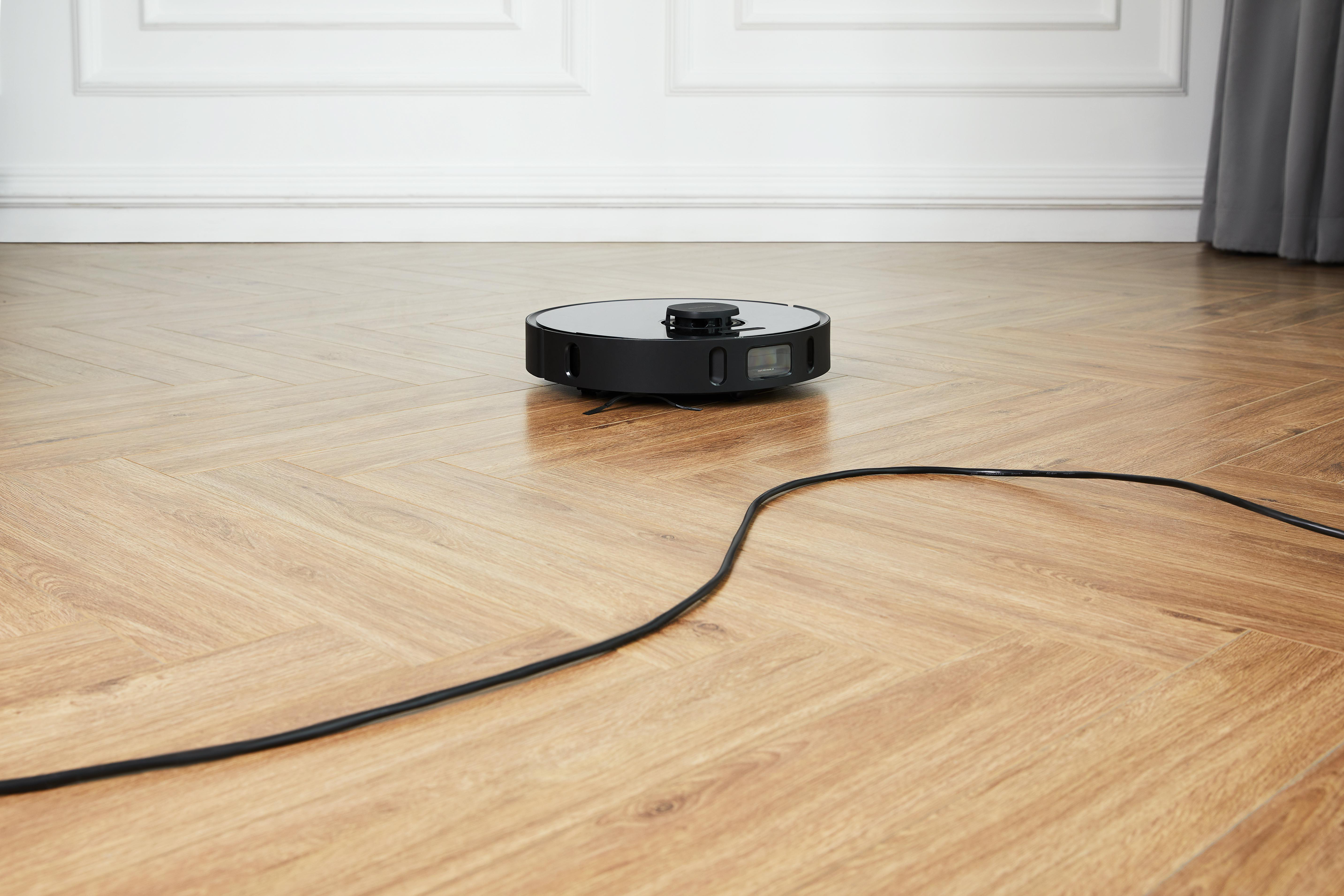 Ein schwarzer Roboter-Staubsauger auf einem Holzboden, das Ladekabel ist sichtbar.