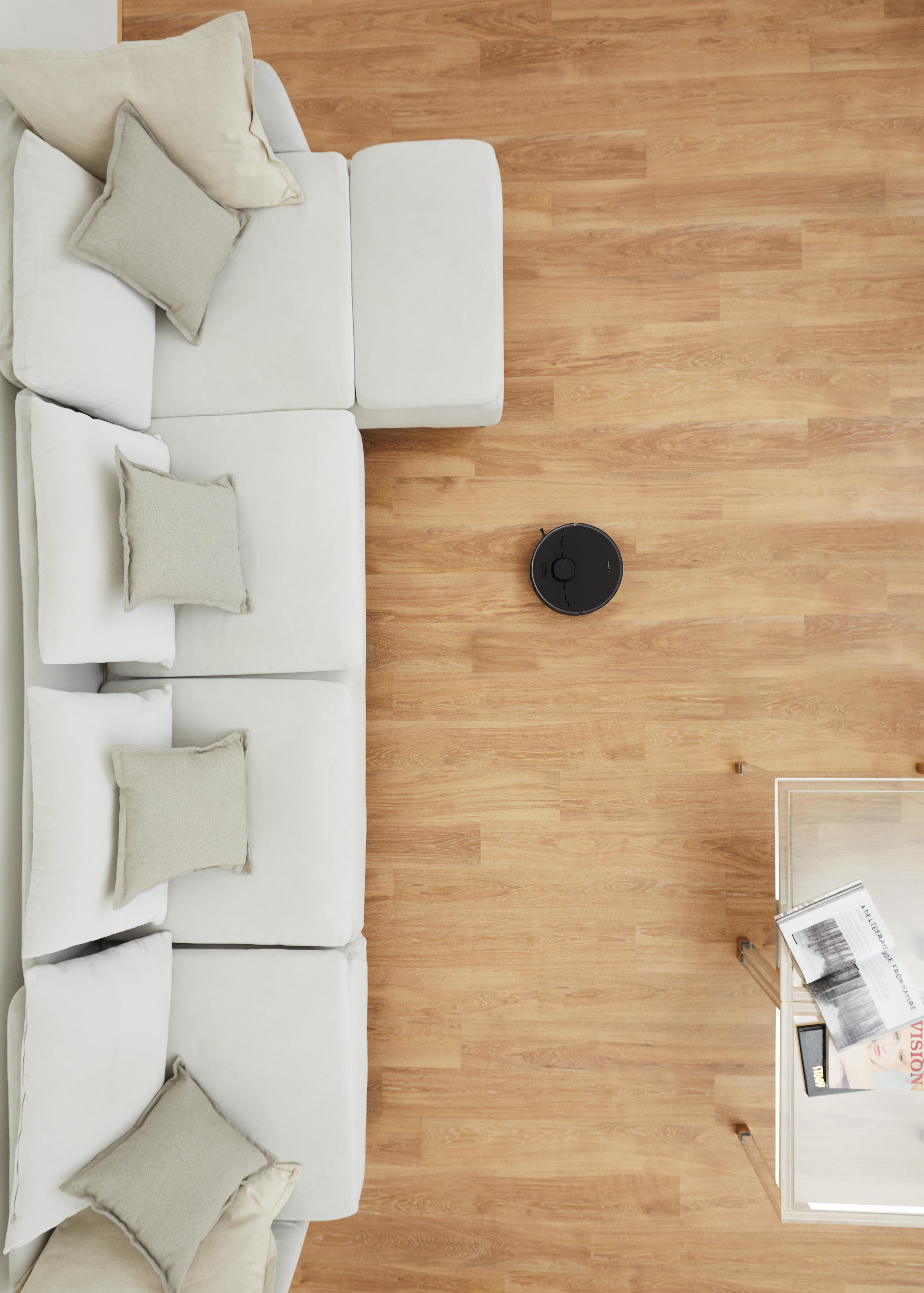 Draufsicht auf ein weißes Sofa mit Kissen, einen Roboterstaubsauger und einen Glastisch.