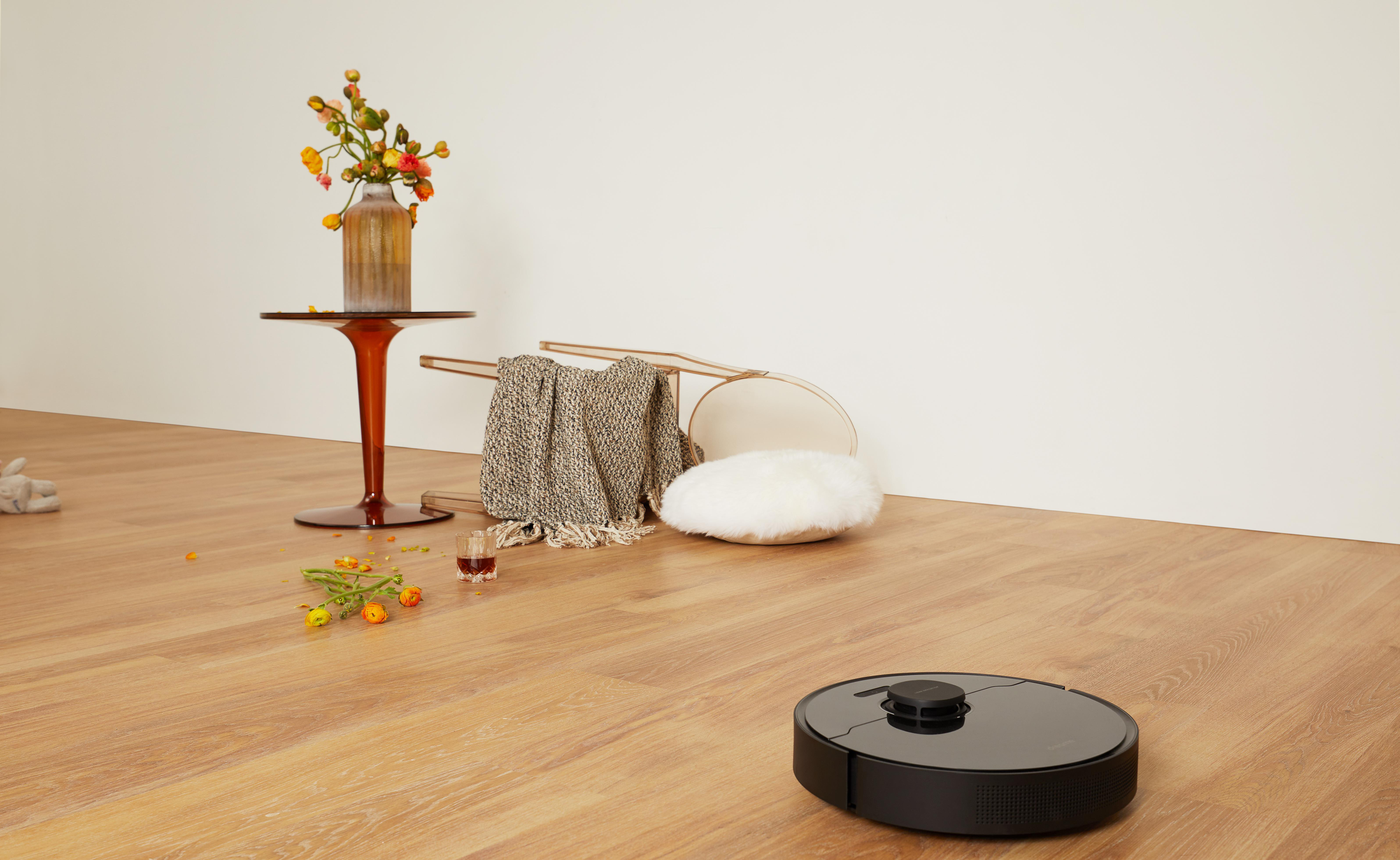 Roboterstaubsauger auf Holzboden, umgekippter Stuhl, Blumen und verstreute Gegenstände.