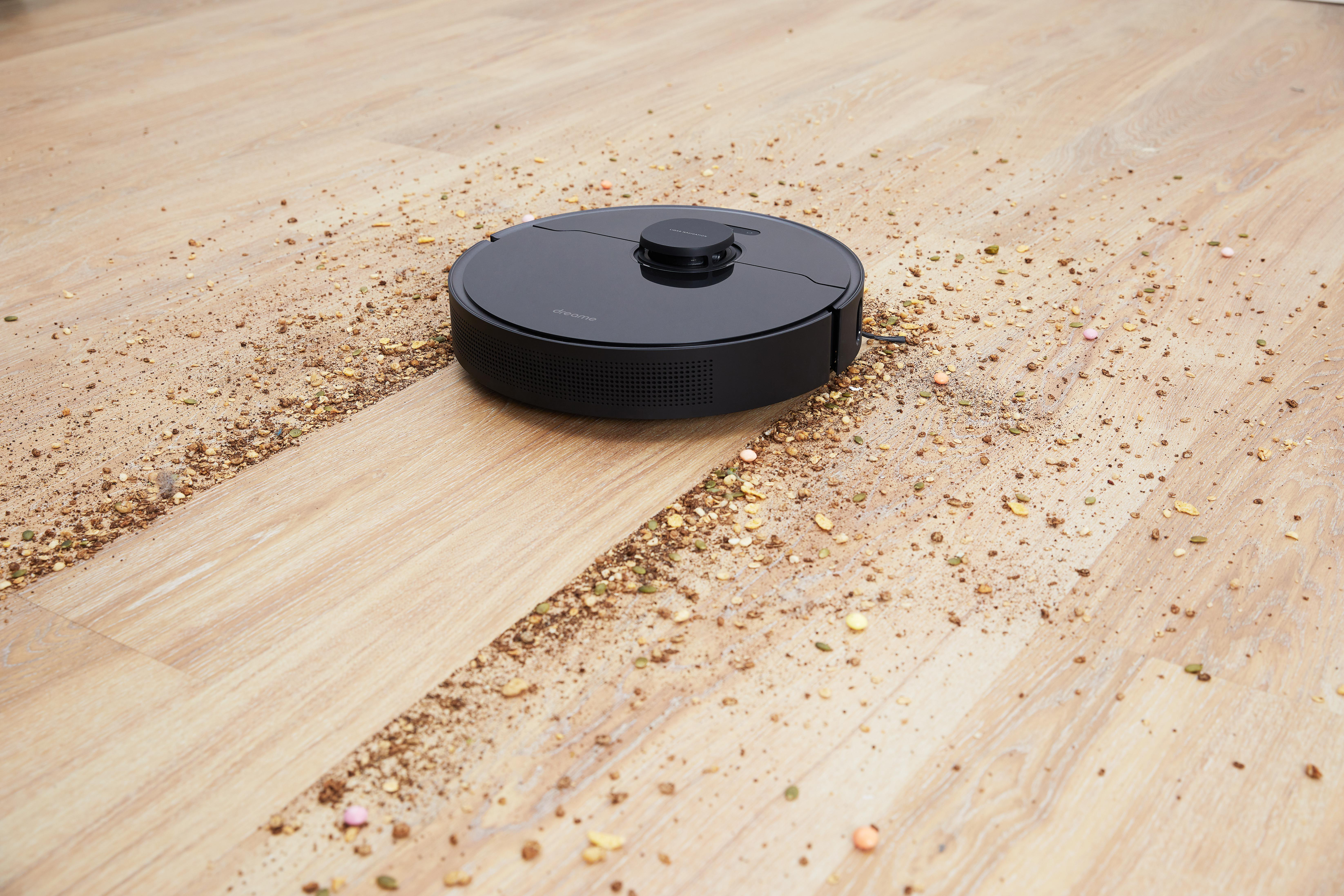 Ein schwarzer Roboter-Staubsauger auf einem Holzboden mit verstreuten Ablagerungen.