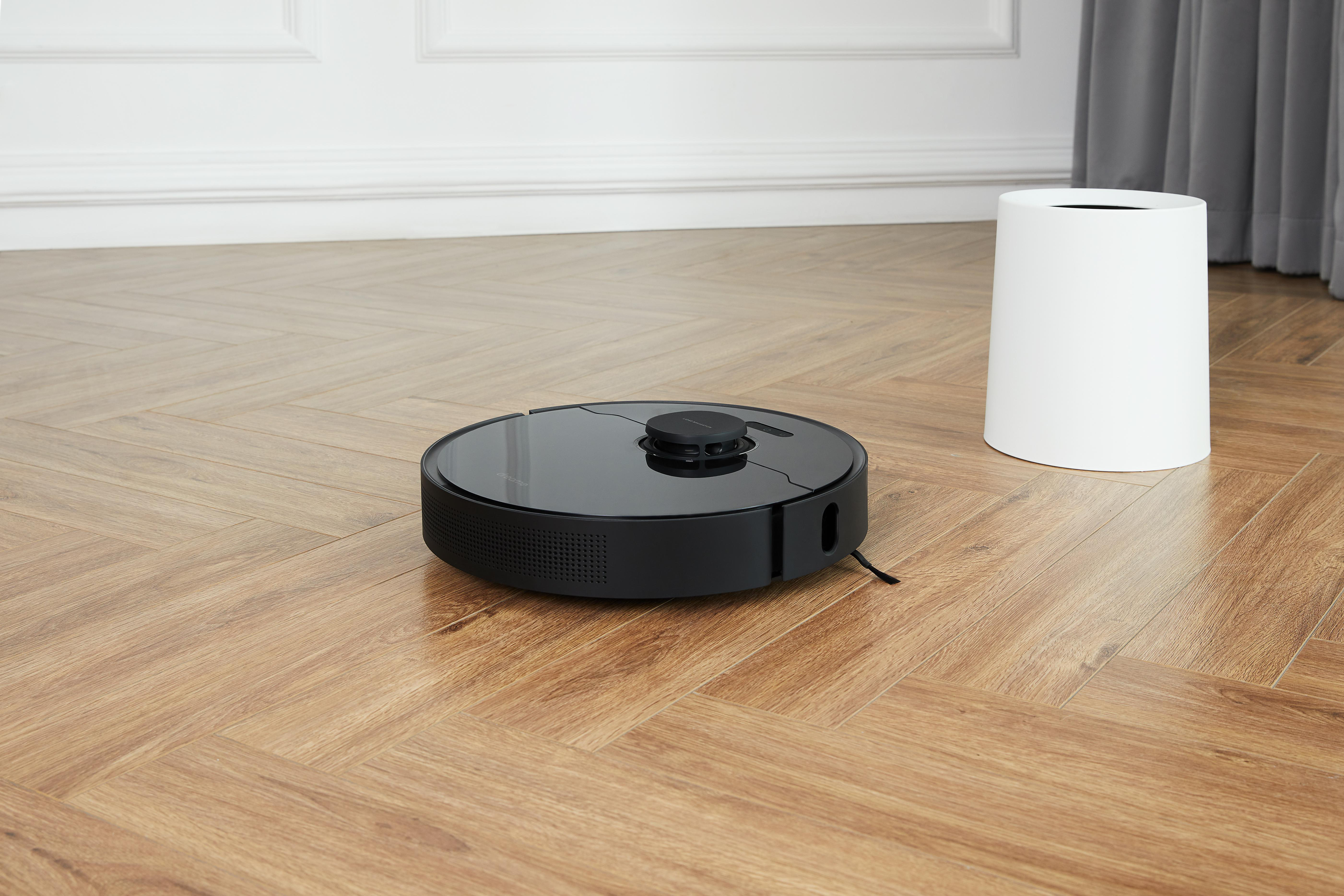 Ein schwarzer Roboter-Staubsauger auf einem Holzboden. Ein weißer zylindrischer Behälter befindet sich im Hintergrund.