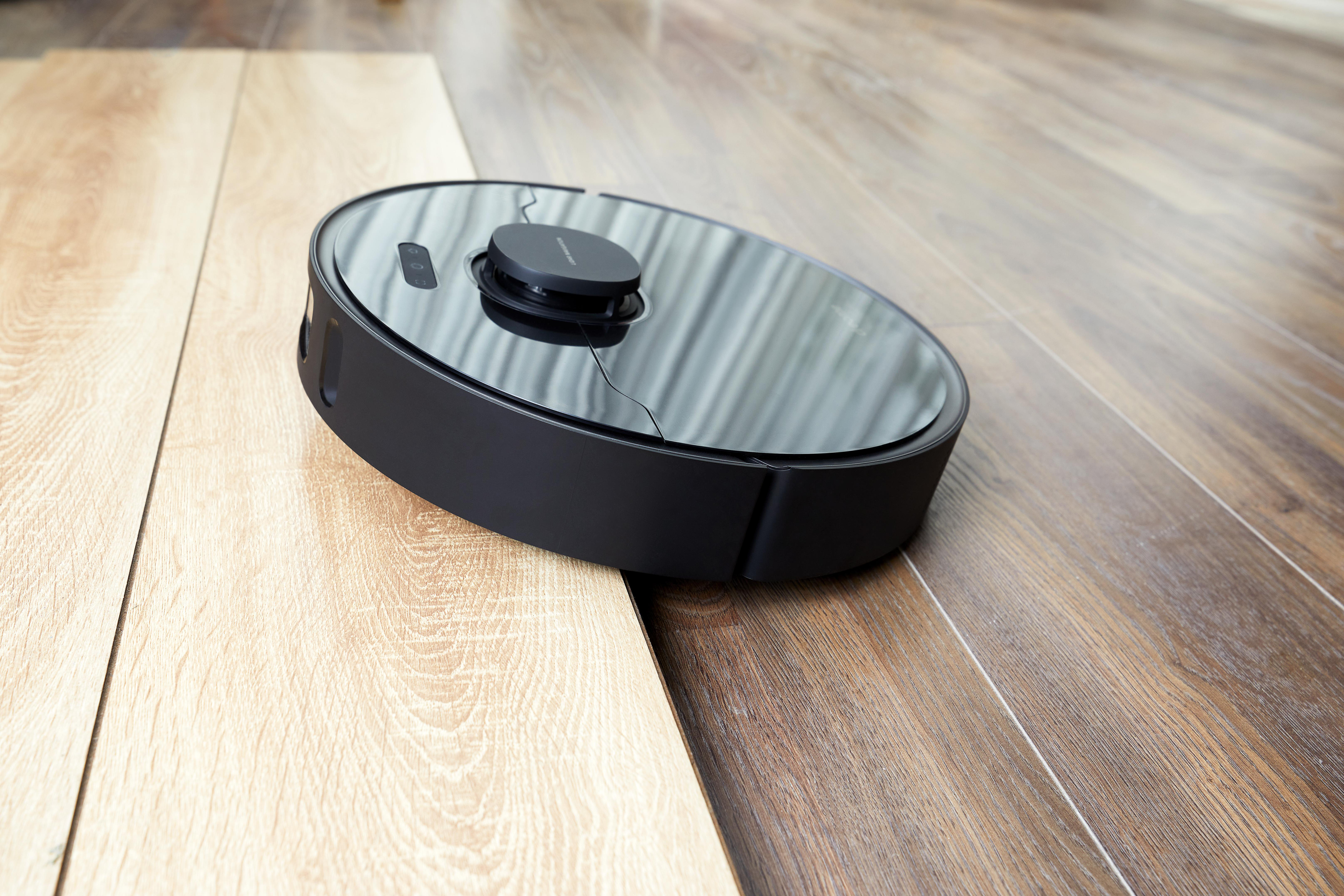 Ein schwarzer Roboter-Staubsauger auf einem Holzboden. Es hat ein kreisförmiges Design mit einem oberen Knopf.