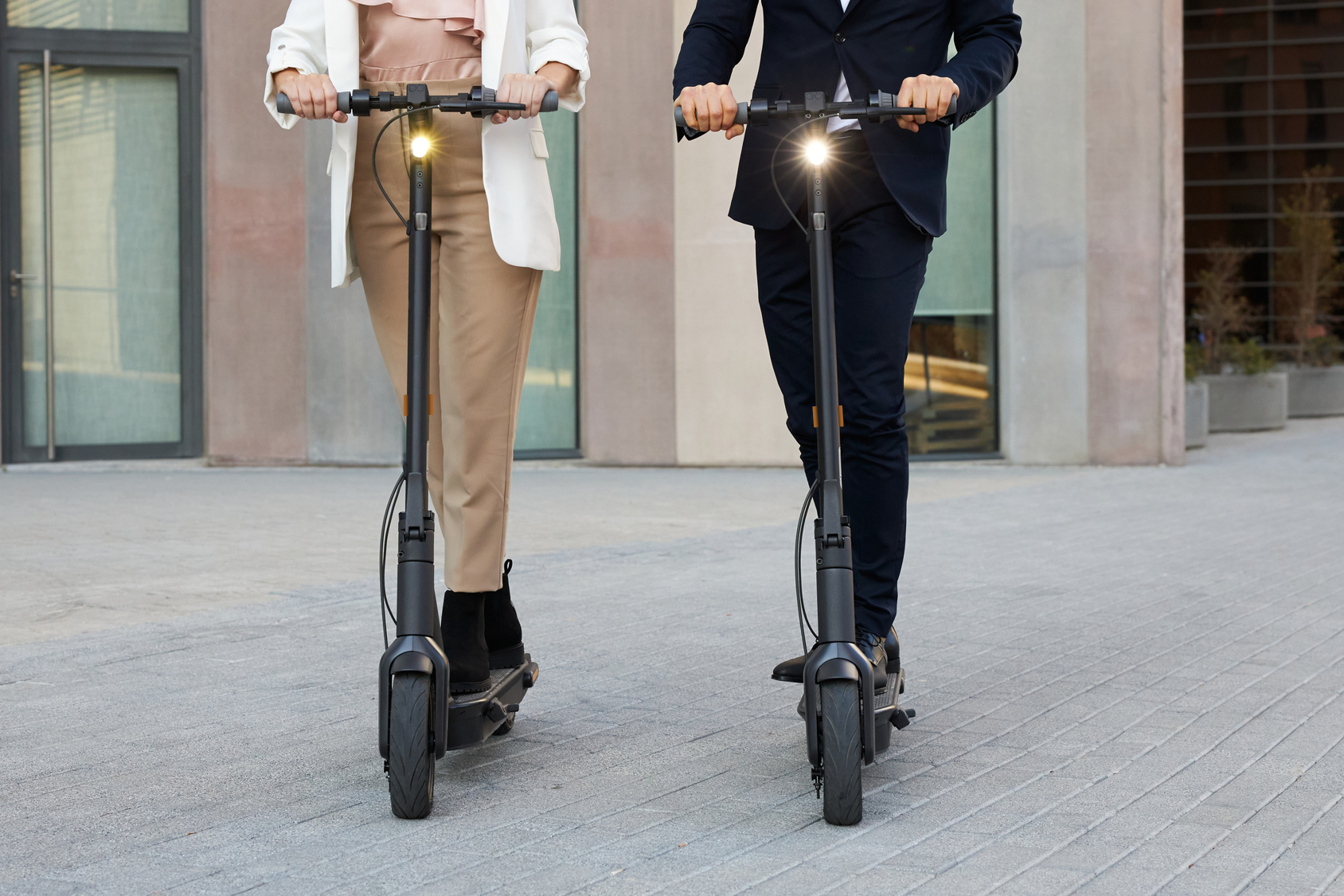 Zwei Personen auf Elektrorollern. Einer trägt einen Blazer, der andere einen Anzug. Sie fahren auf einem Weg.