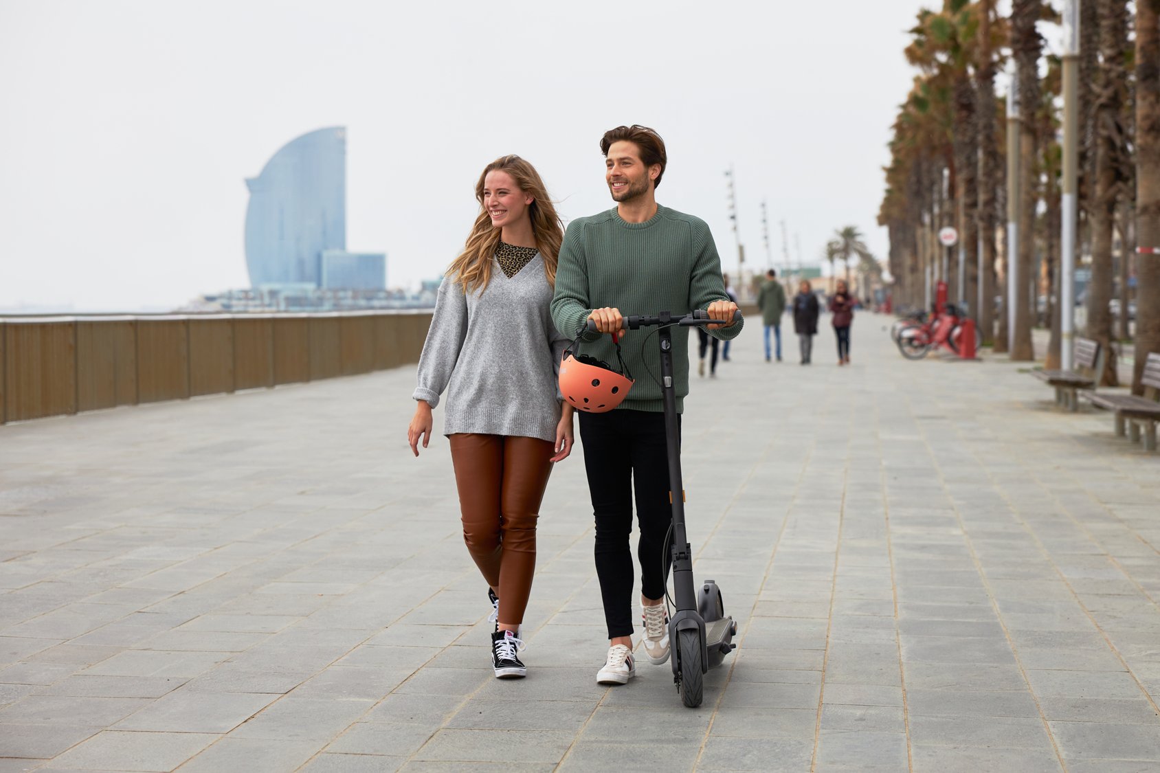Ein Paar geht und der Mann schiebt einen Elektroroller auf einer Promenade mit Stadtkulisse.