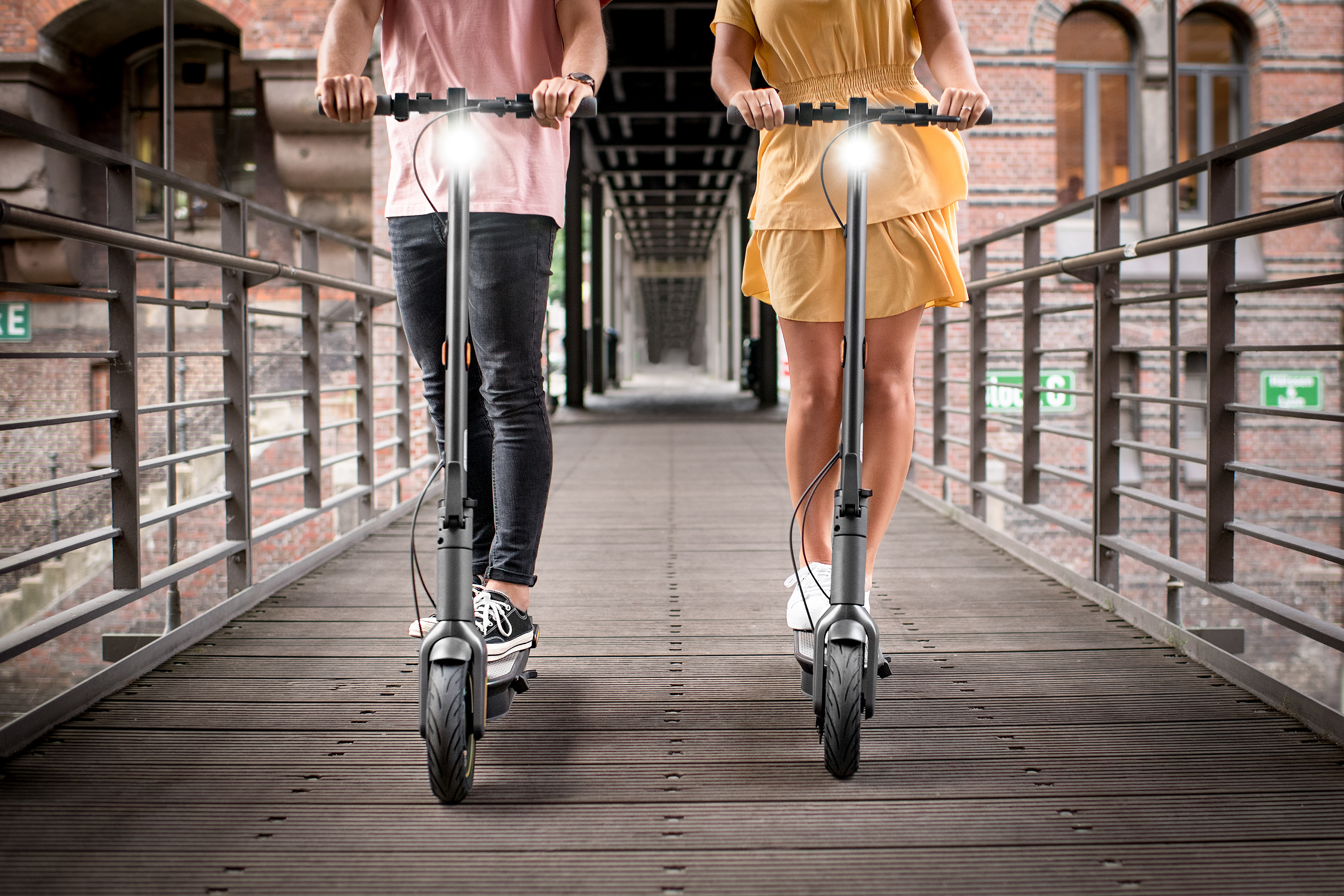 Zwei Personen fahren nebeneinander auf einer Holzbrücke Roller.