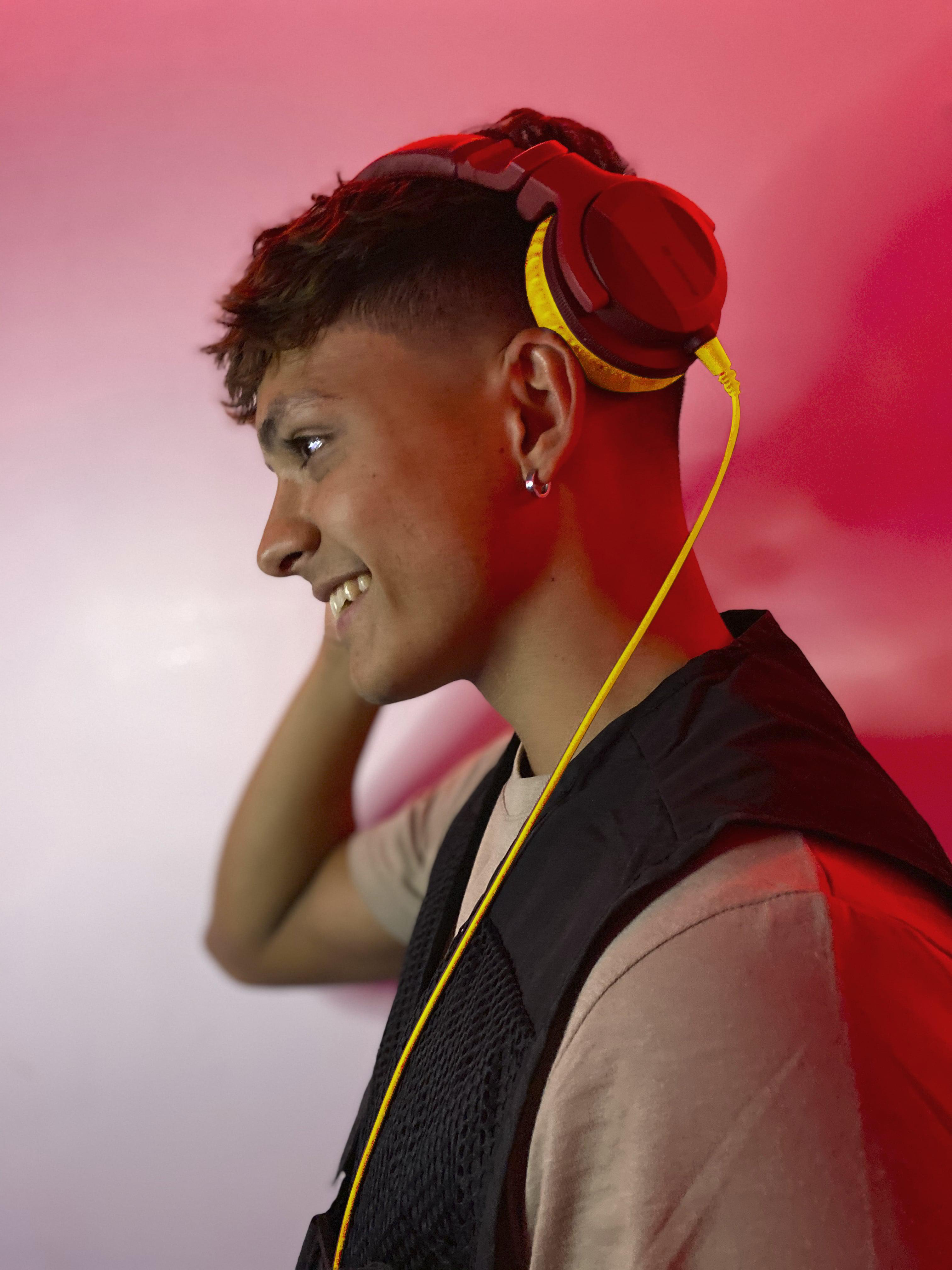 Junger Mann mit Kopfhörern, lächelnd, in einem Studio mit rotem und pinkfarbenem Hintergrund. Weste.