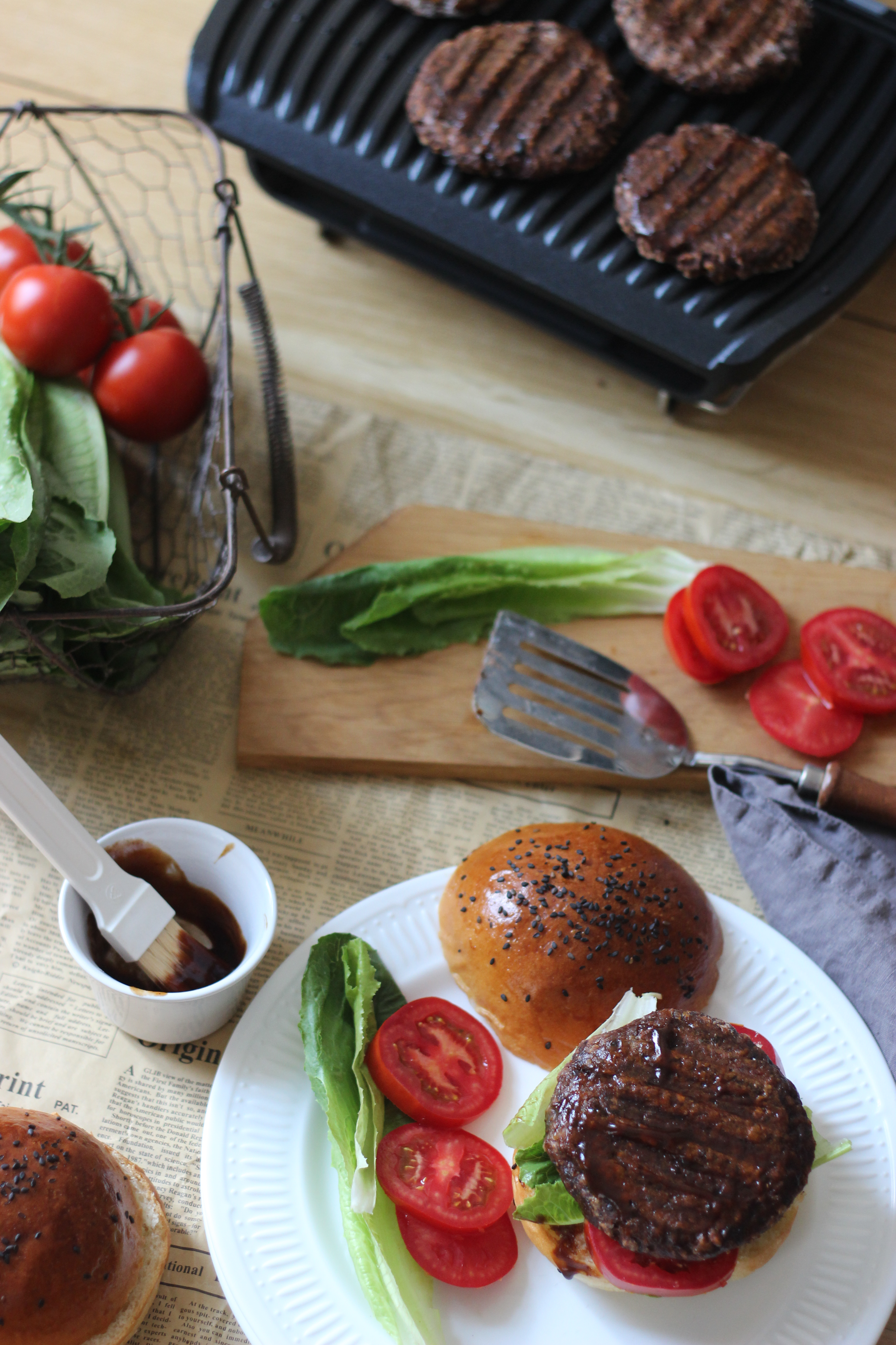 Burger tányéron paradicsommal és salátával. Összetevők az asztalon, grill a hamburgerekkel.
