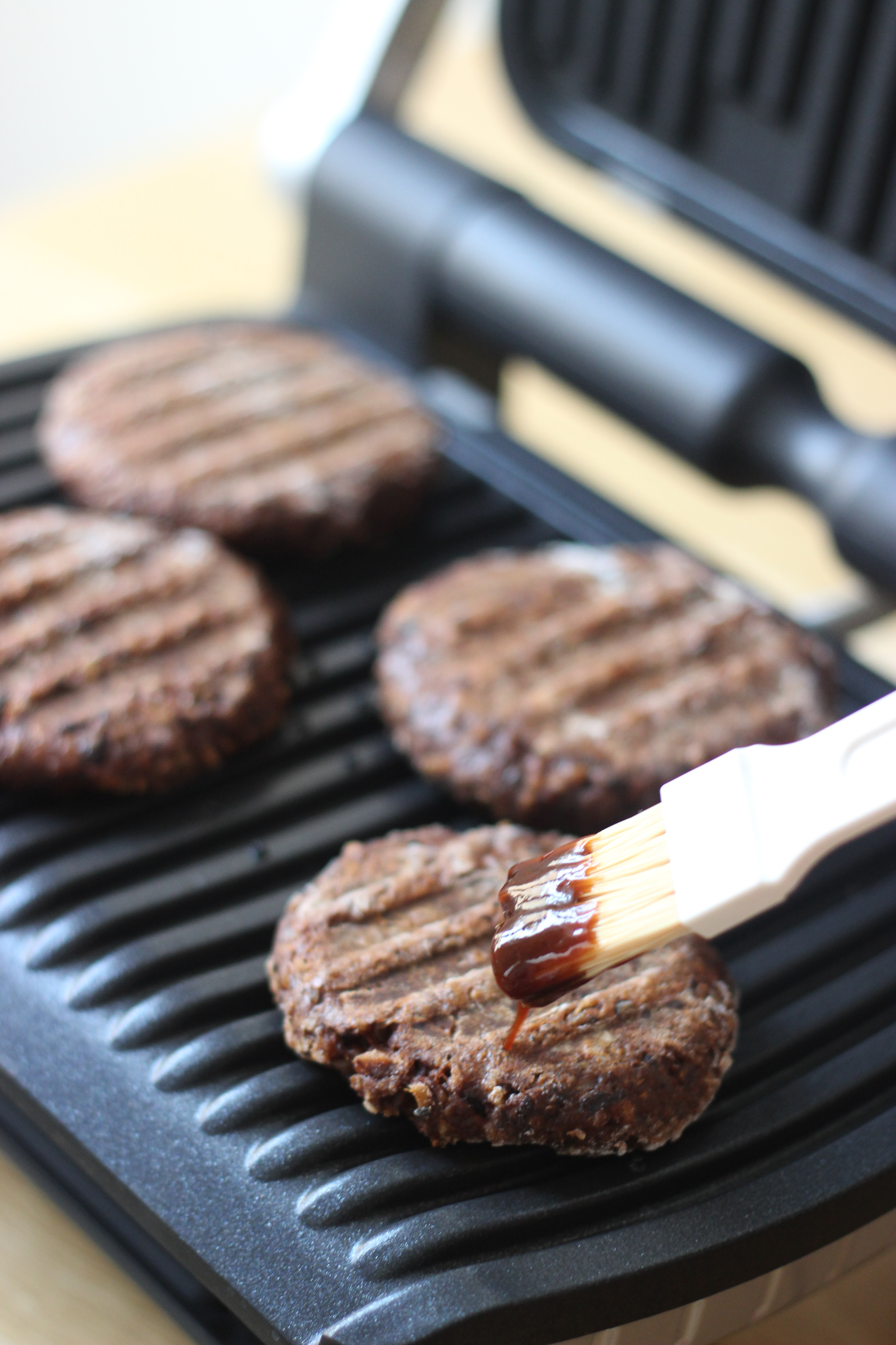 Négy grillezett burger egy fekete grillen. Egy ecset mártást visz fel az egyik burgerre.