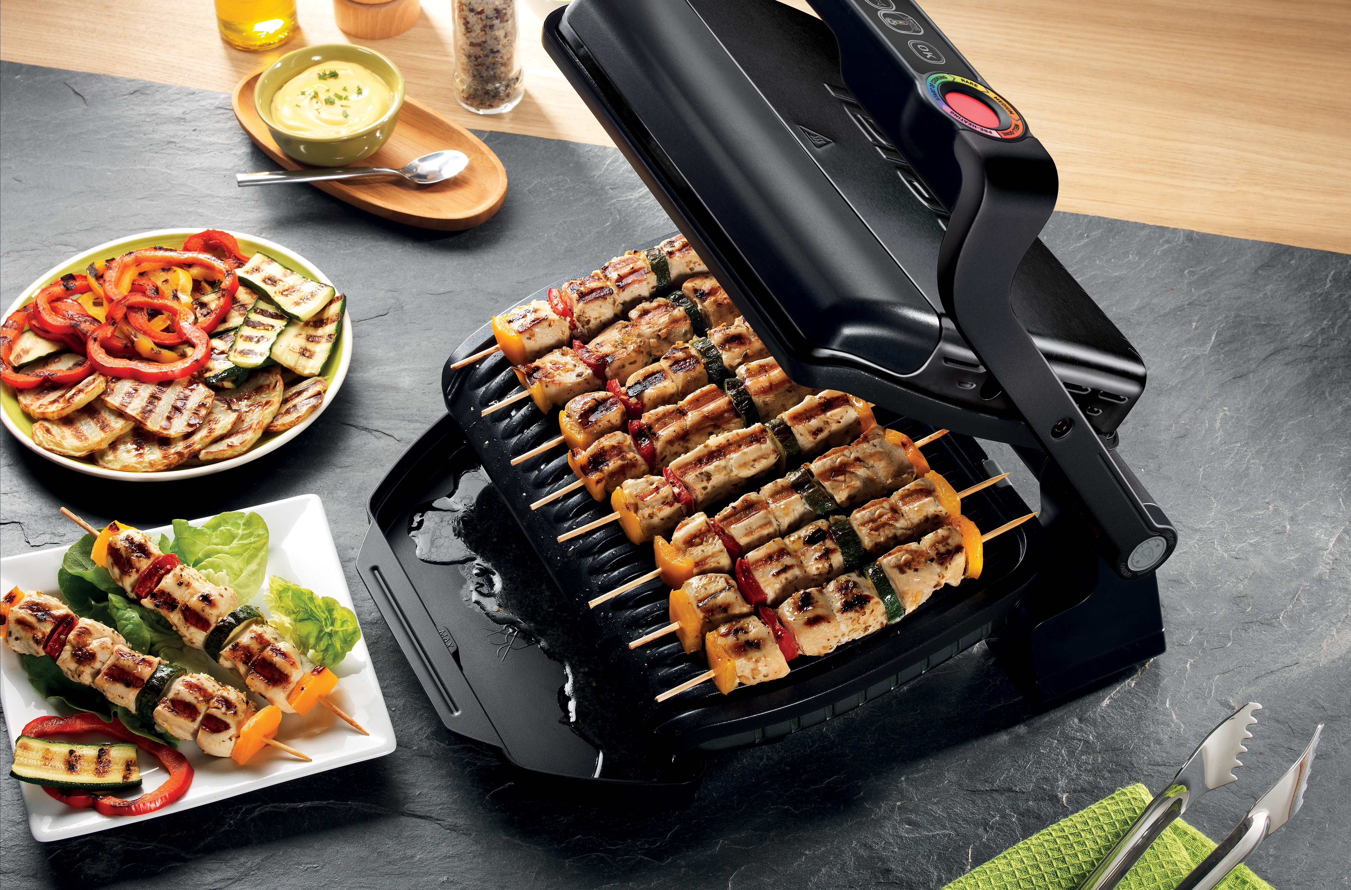 Egy grill grillezett nyársakkal és zöldségekkel, köretekkel és evőeszközökkel egy asztalon.