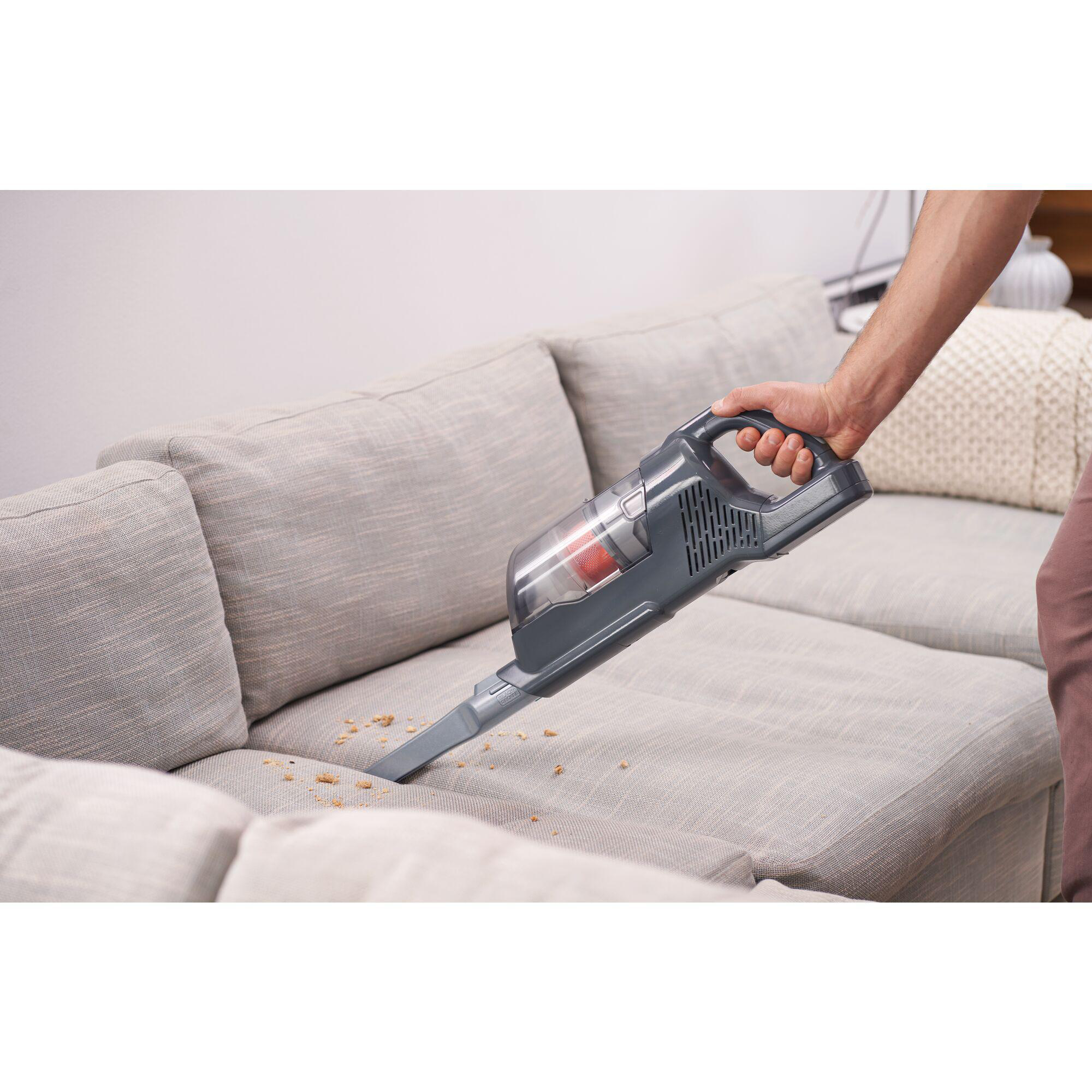 Eine Person benutzt einen grauen kabellosen Staubsauger, um ein graues Sofa zu reinigen, mit sichtbaren Krümeln.