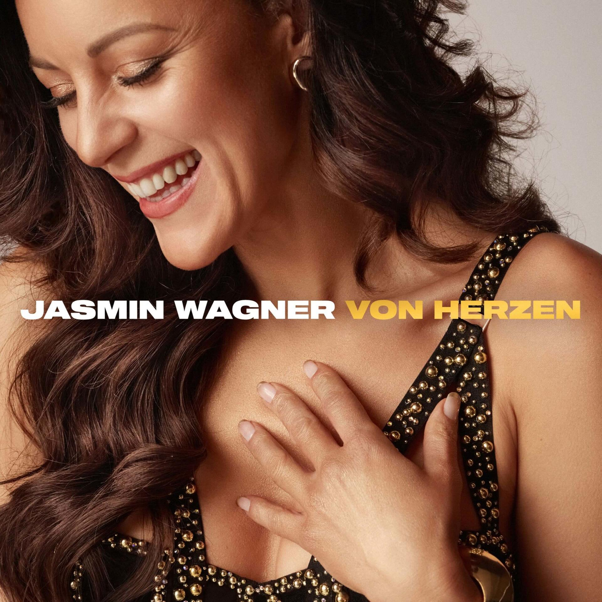 Mosolygó nő, kezét a mellén, fekete ruha arany szegecsekkel. A szöveg: 'Jasmin Wagner von Herzen'.