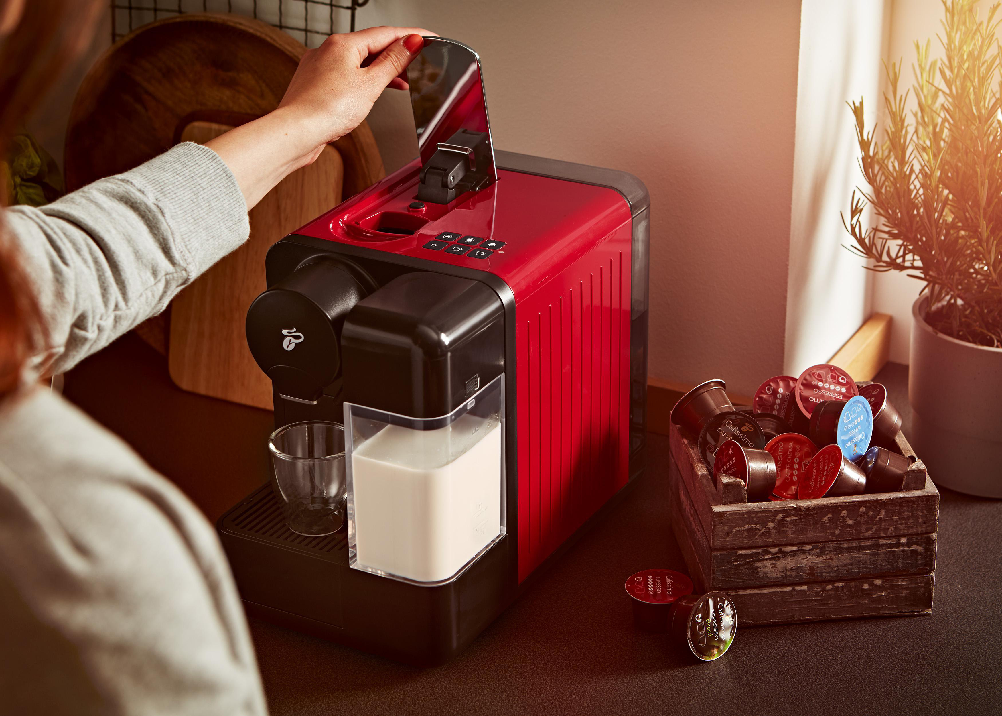 Person bedient eine rote Kaffeemaschine. Kaffeemaschine neben Holzkiste mit Kapseln.