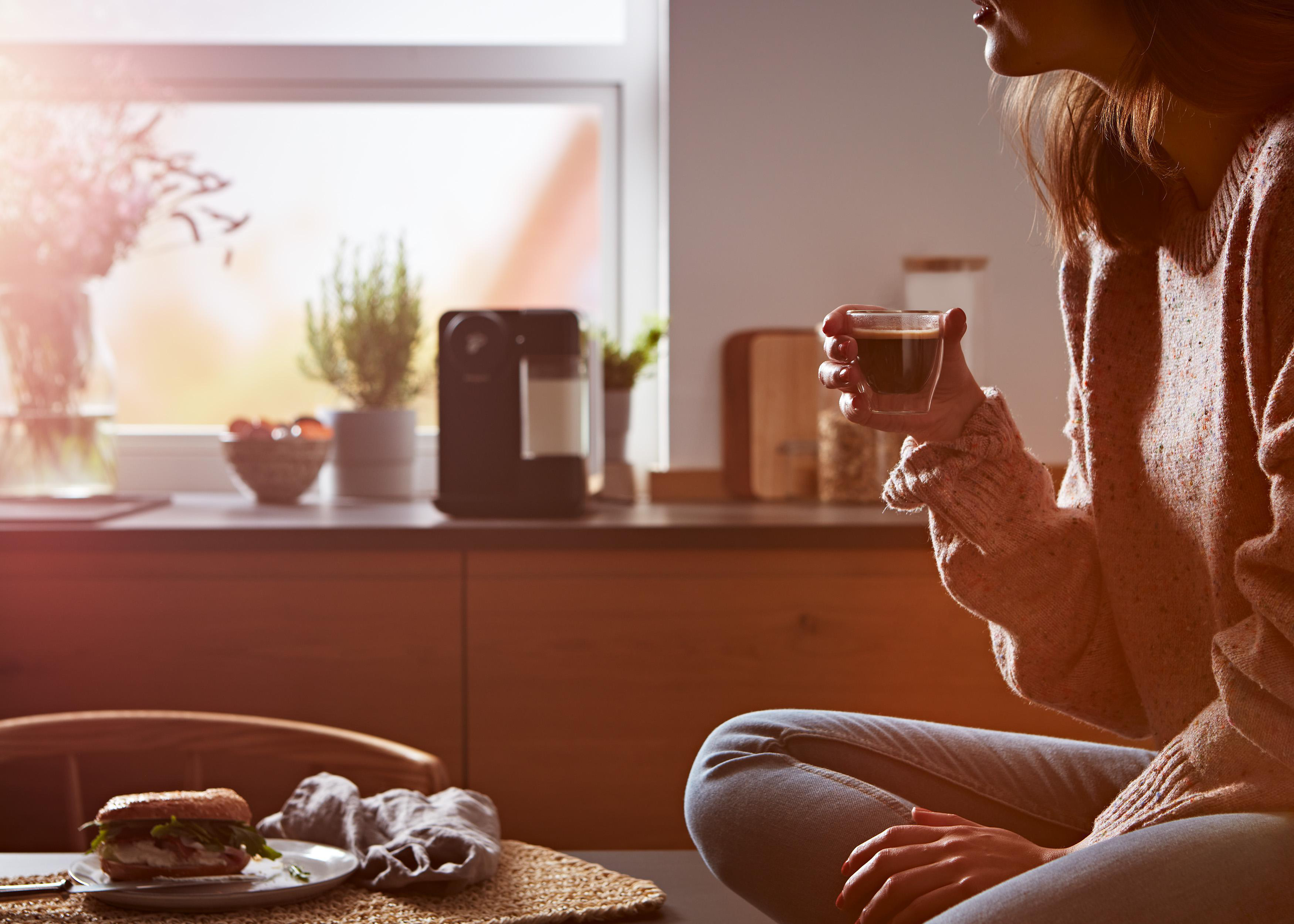 Eine Person hält eine Tasse Kaffee, mit einem Sandwich und einer Kaffeemaschine im Hintergrund.