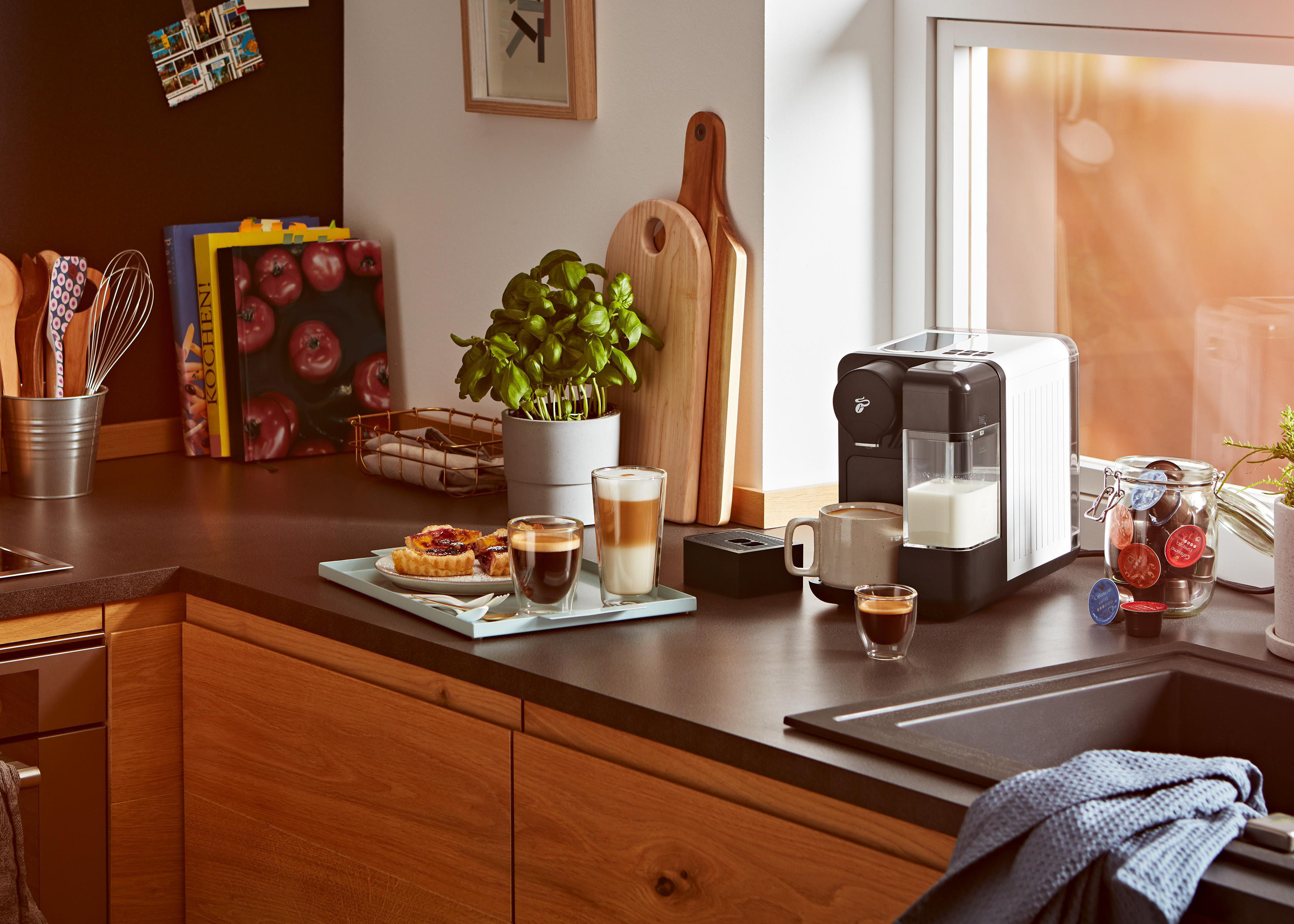 Küchentheke mit Kaffeemaschine, Getränken und Zutaten. Holzschränke und Fenster im Hintergrund.