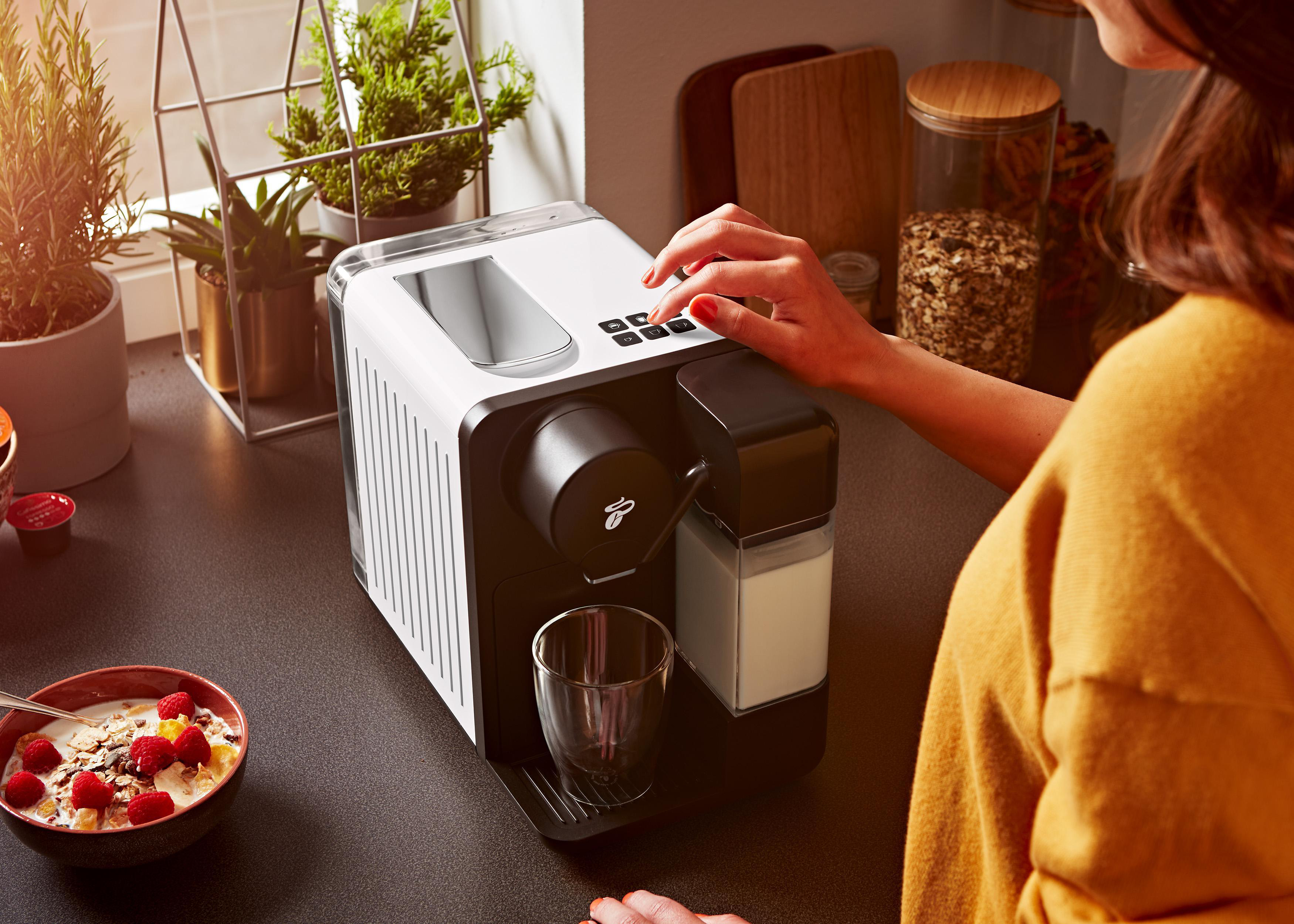 Frau bedient eine schwarz-weiße Kaffeemaschine. Eine Schüssel mit Müsli im Vordergrund.