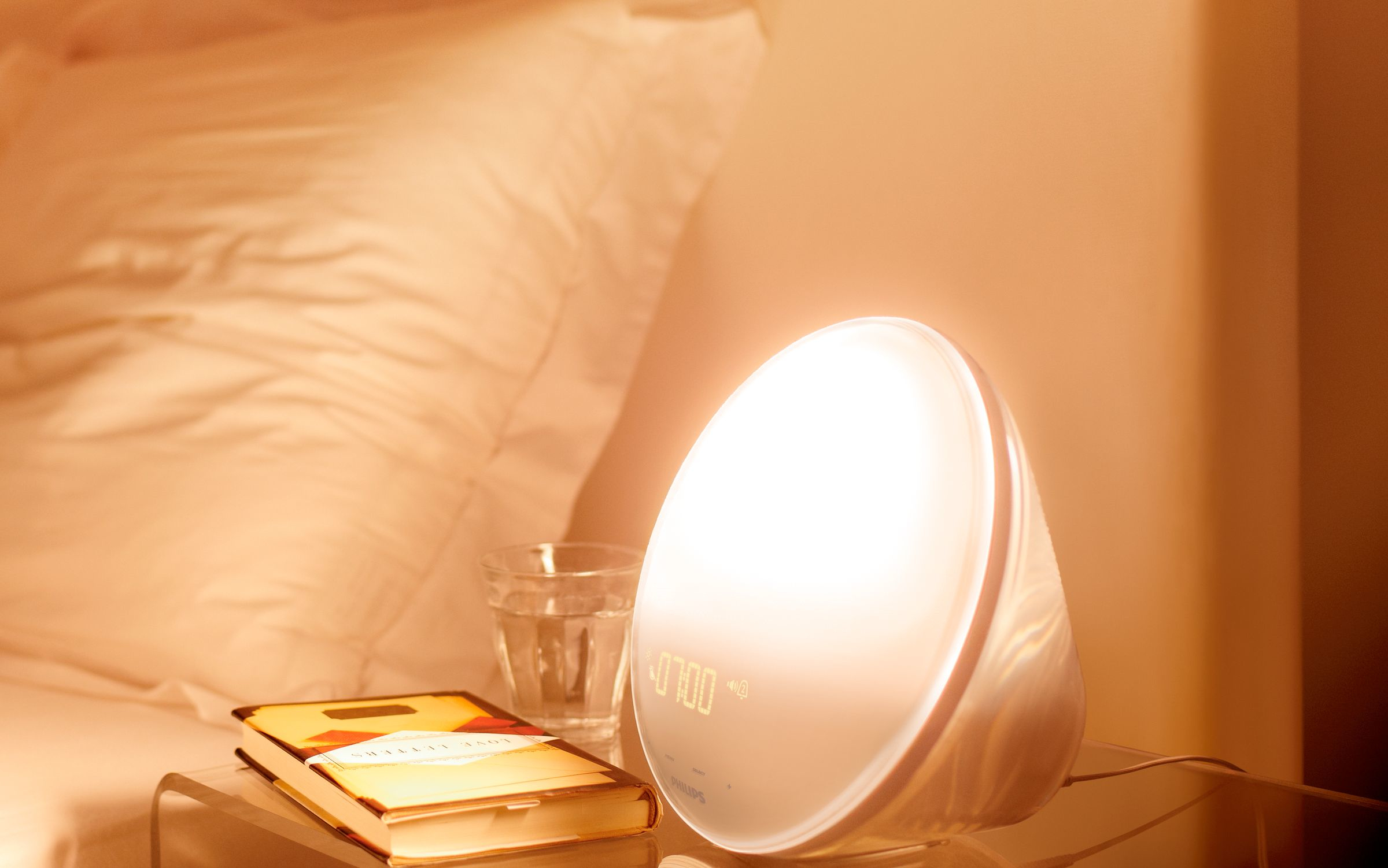 Lampe de réveil Philips blanche avec l'heure 07h00 affichée, à côté d'un livre et d'un verre.