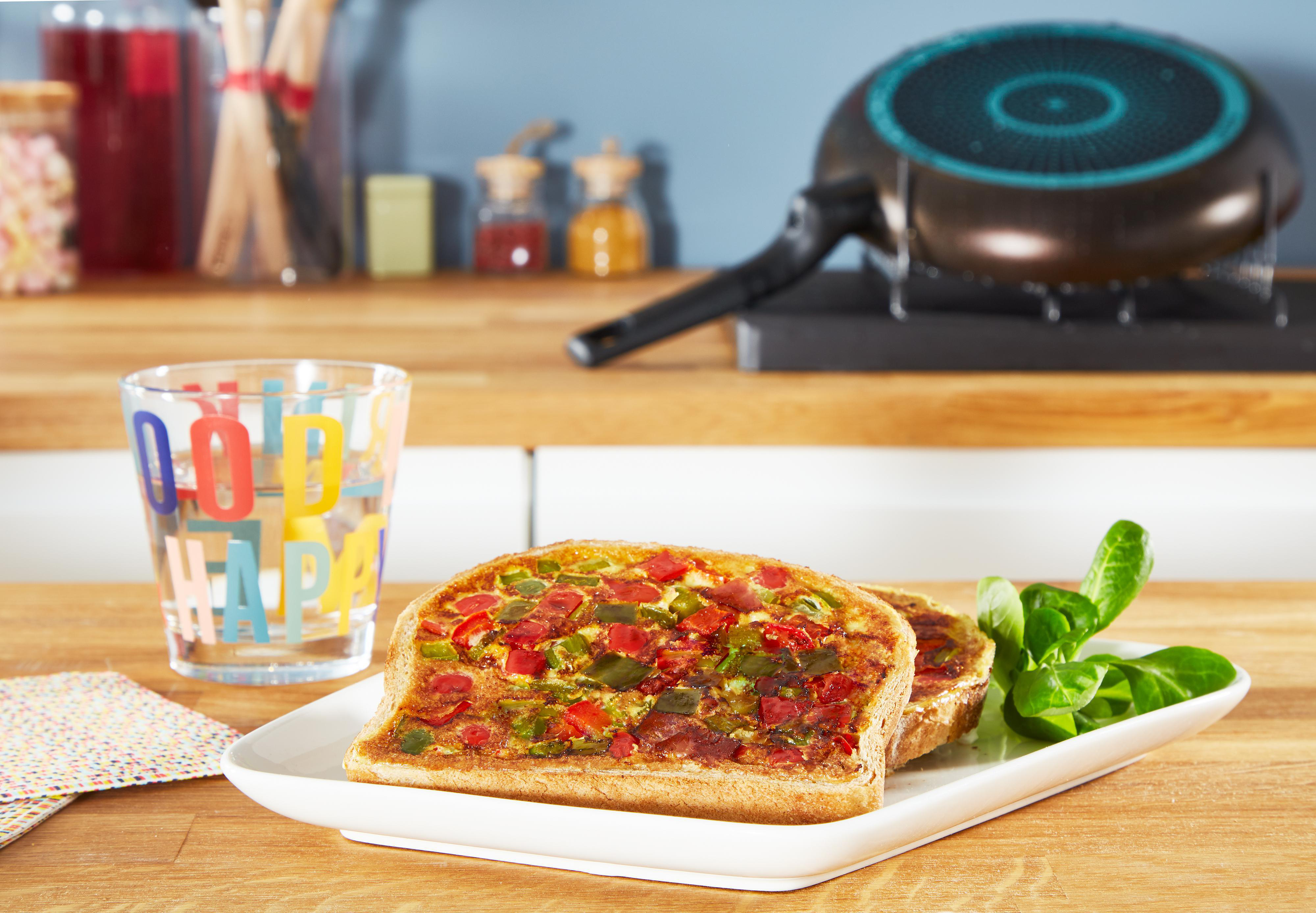 Zwei Toastscheiben mit gekochtem Gemüse auf einem weißen Teller, mit einem Glas und einer Pfanne.