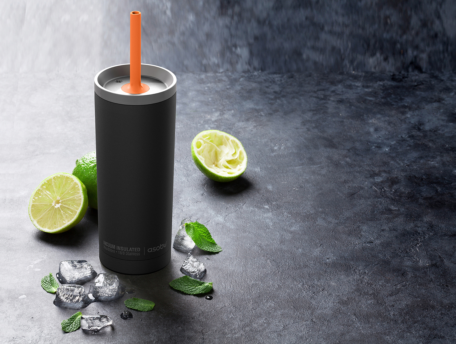 A black insulated tumbler with an orange straw, limes, ice cubes, and mint leaves on a surface.
