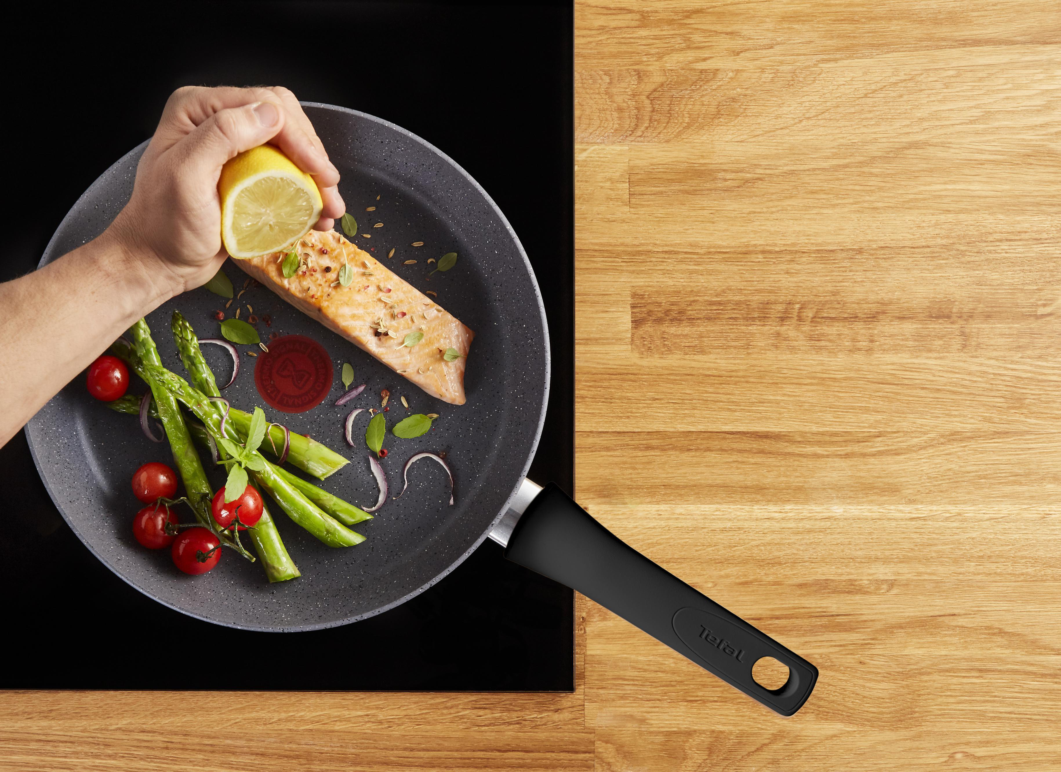 Eine Person gibt Zitronensaft zu Lachs in einer Bratpfanne mit Spargel und Tomaten auf Holzoberfläche.