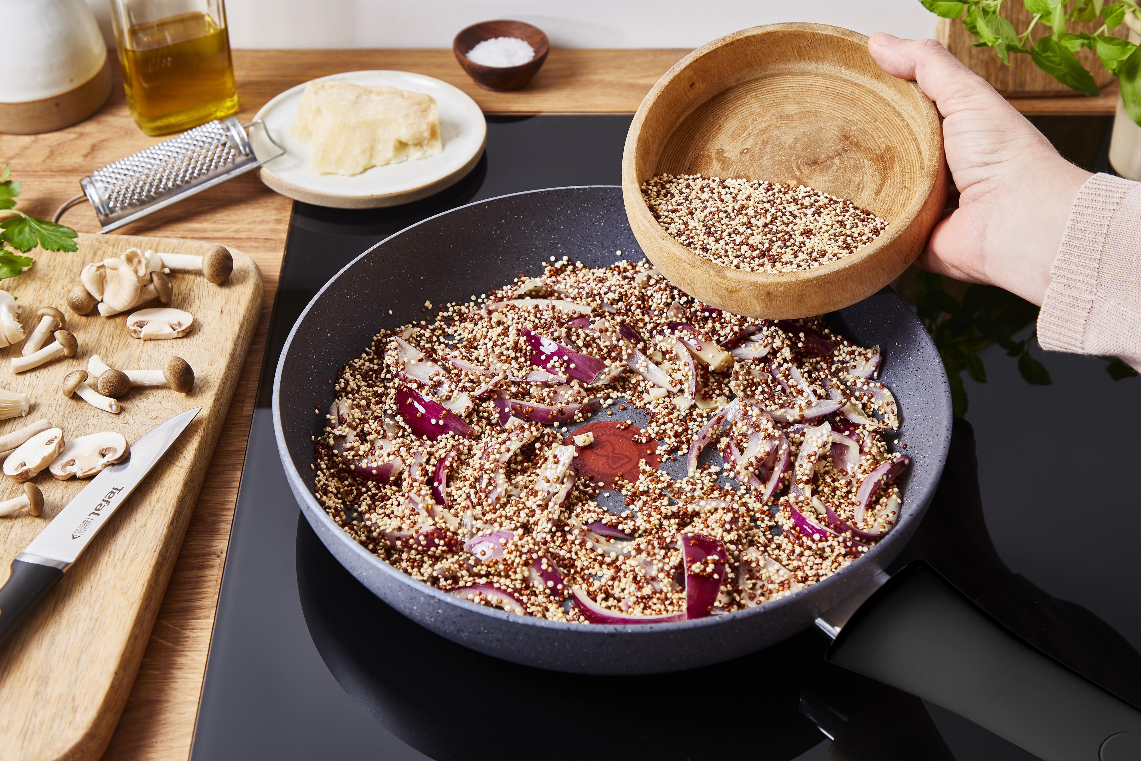 Eine Pfanne mit Essen, das gekocht wird. Eine Holzschüssel gießt Quinoa in die Pfanne mit roten Zwiebeln.
