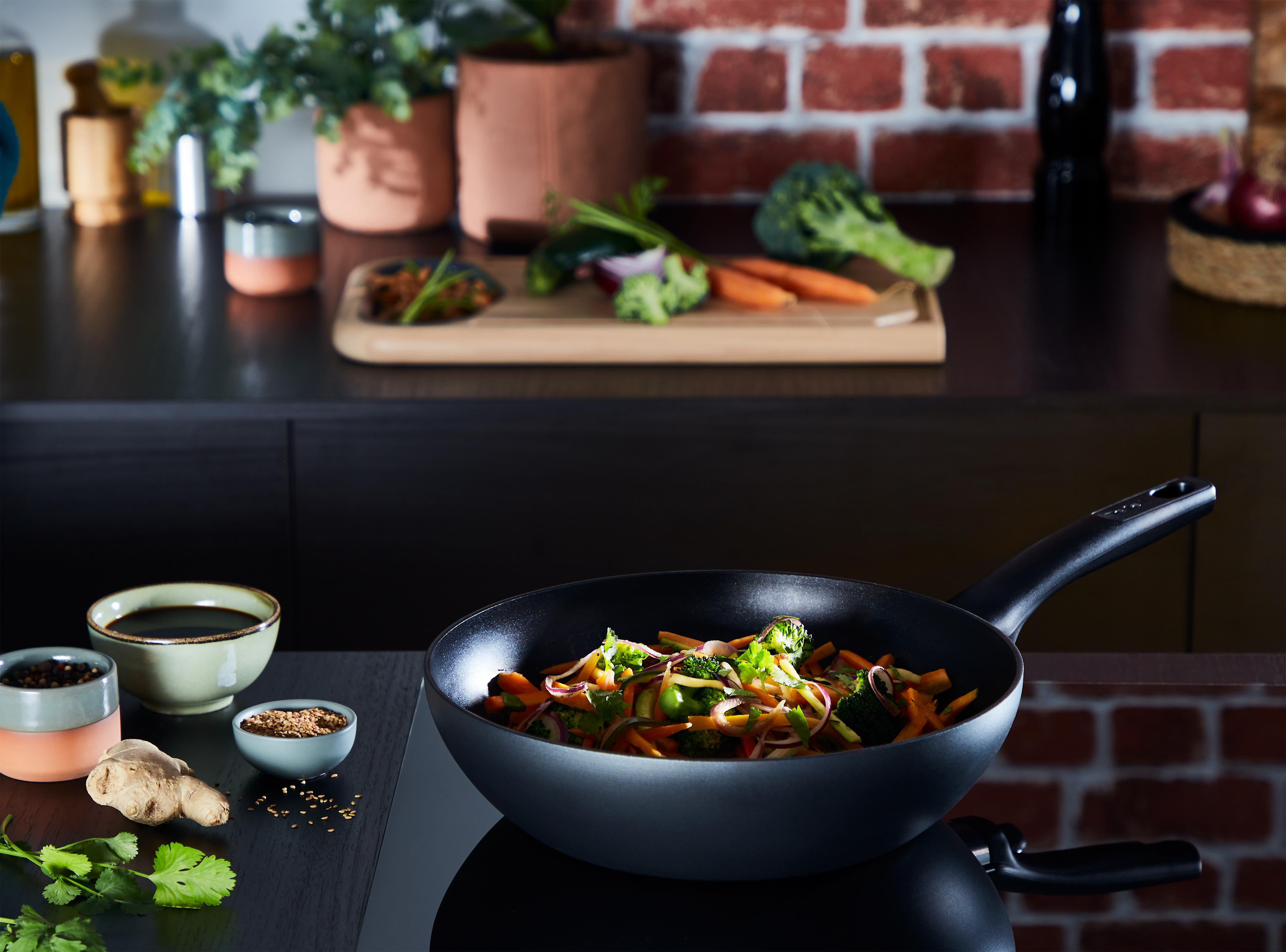 Wok-Pfanne mit Gemüse im Zentrum, umgeben von Zutaten auf der Küchentheke.