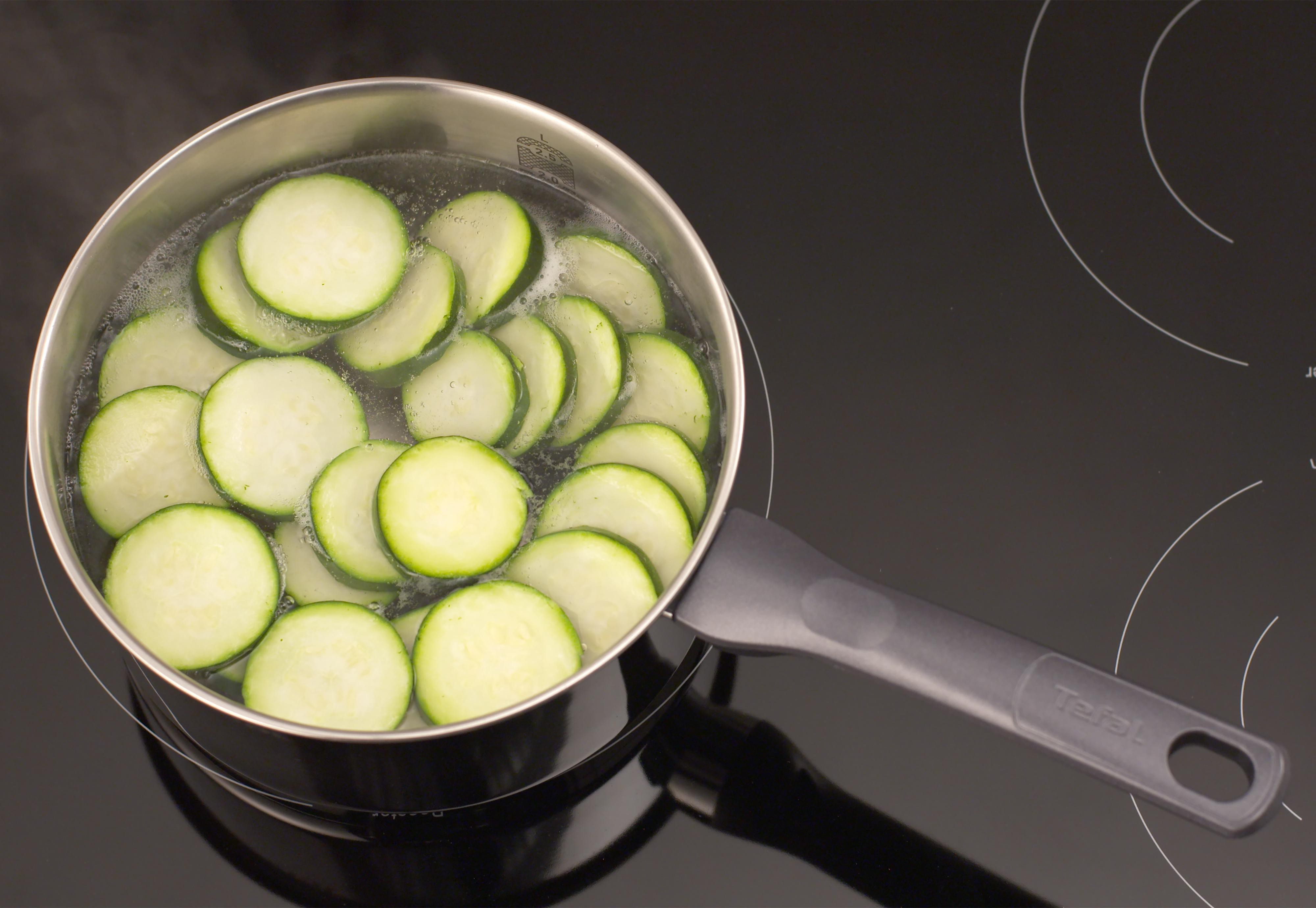 In Scheiben geschnittene Zucchini, die in einem Edelstahltopf auf einem schwarzen Kochfeld kochen.