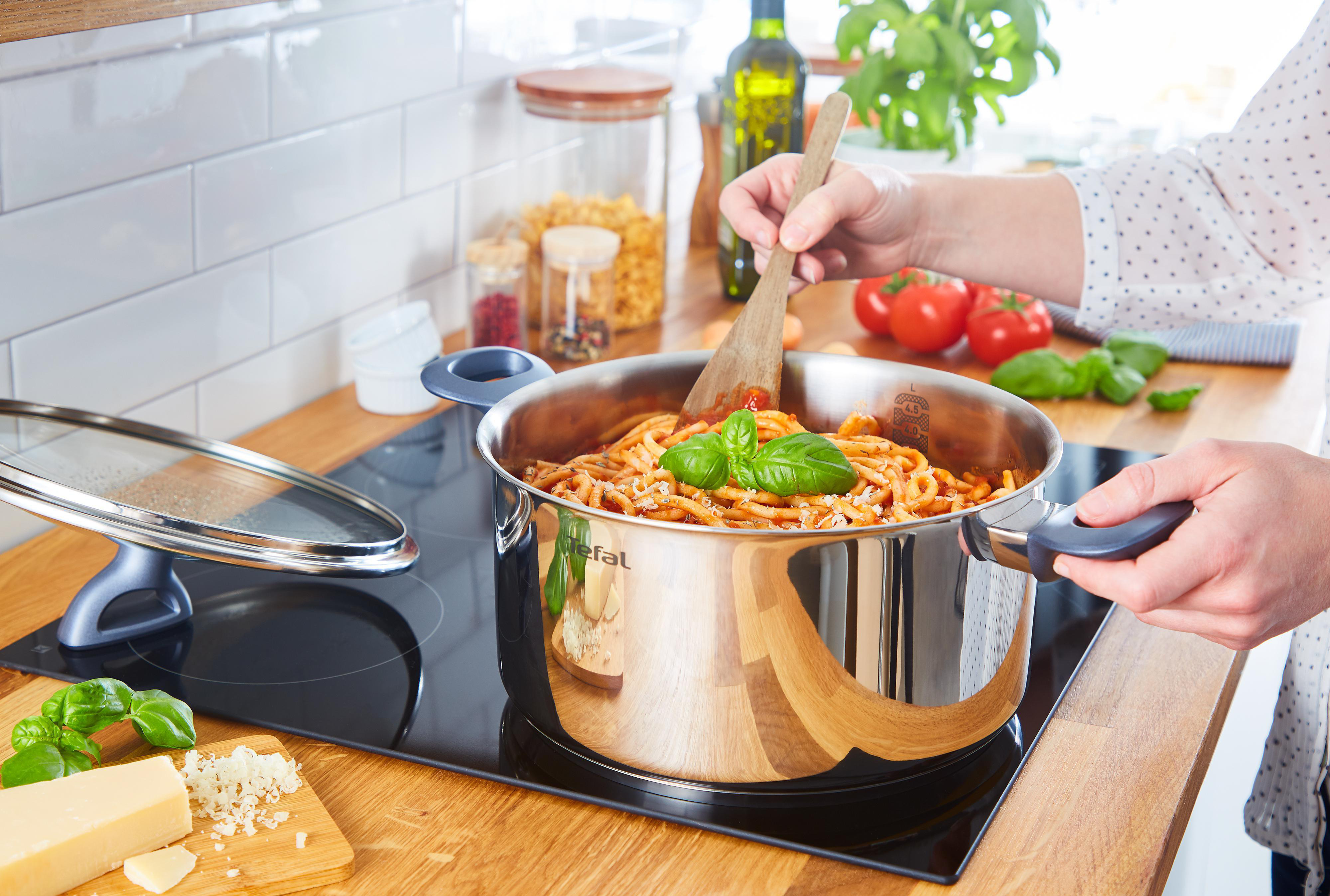 Eine Person rührt Pasta in einem Topf auf einem Herd um. Zutaten wie Tomaten und Basilikum sind in der Nähe.