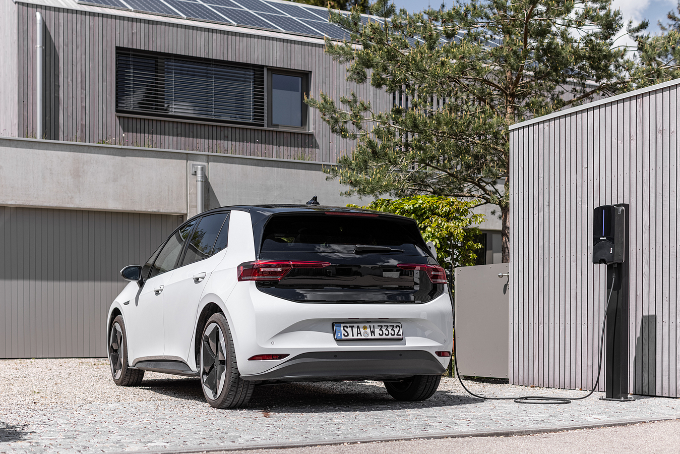 Weißes Elektroauto vor einem Haus, das geladen wird. Das Haus hat Sonnenkollektoren auf dem Dach.