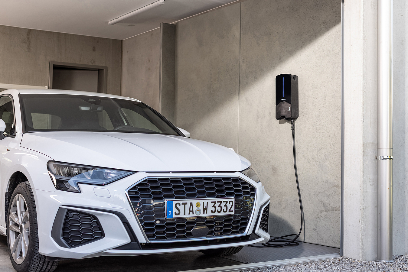 Ein weißes Auto steht in der Nähe einer Ladestation. Ein schwarzes Ladegerät ist an der Wand montiert.
