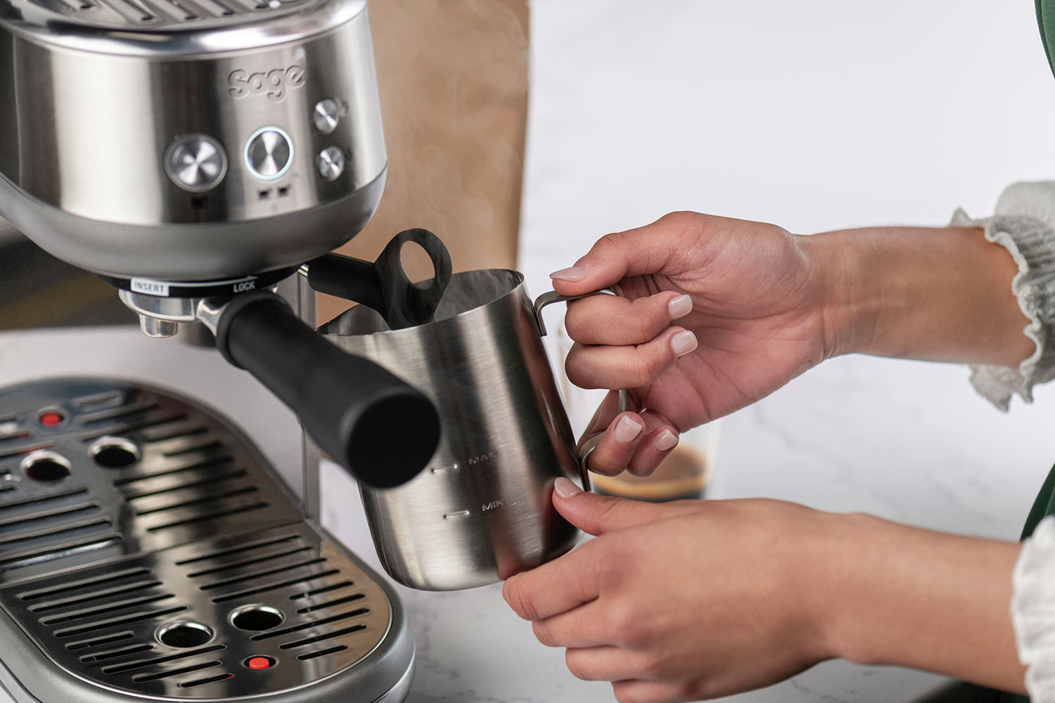 A silver espresso machine with a person pouring milk from a steel pitcher. A brown paper bag is in the background.