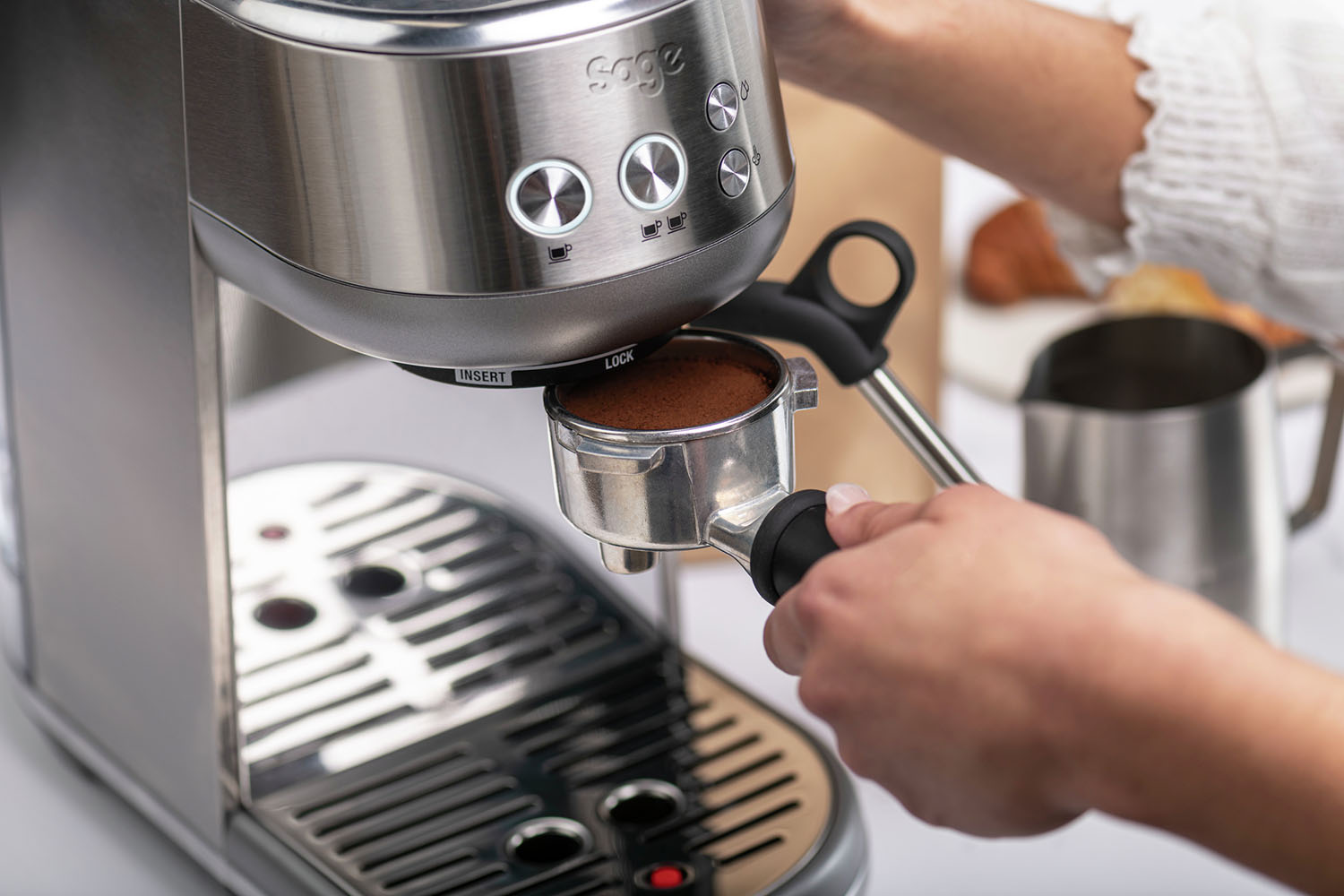 Person holding a portafilter with coffee grounds, near a silver espresso machine. Background is white.