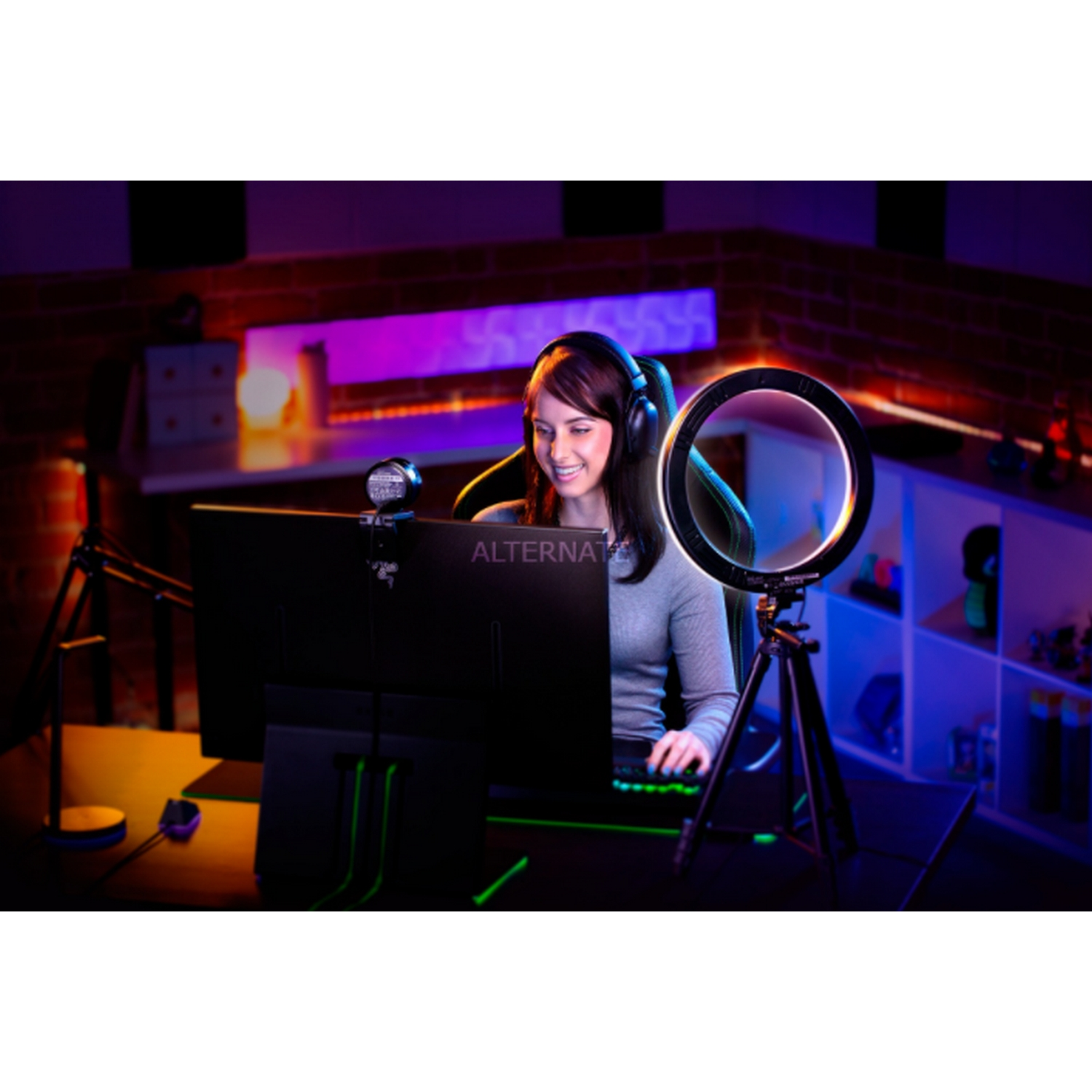 Mujer con auriculares frente a un monitor, iluminada por una luz de anillo. Teclado y escritorio visibles.