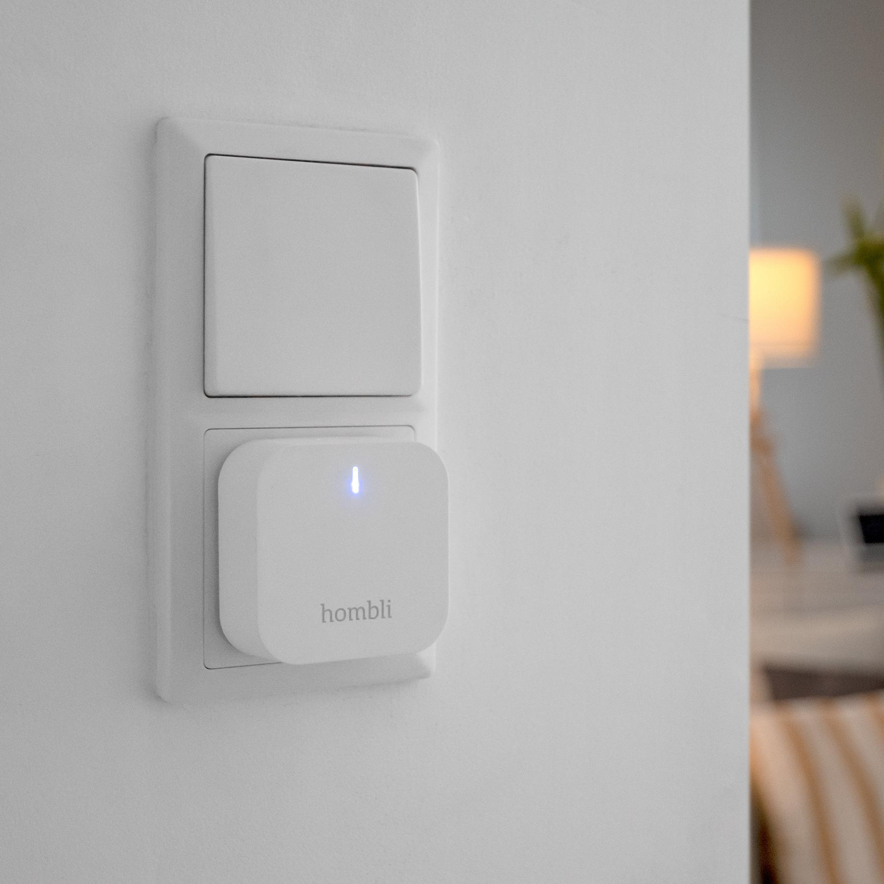 Eine weiße Wand mit einem weißen Lichtschalter und einem weißen Smart Plug mit blauem Licht. Raum im Hintergrund.