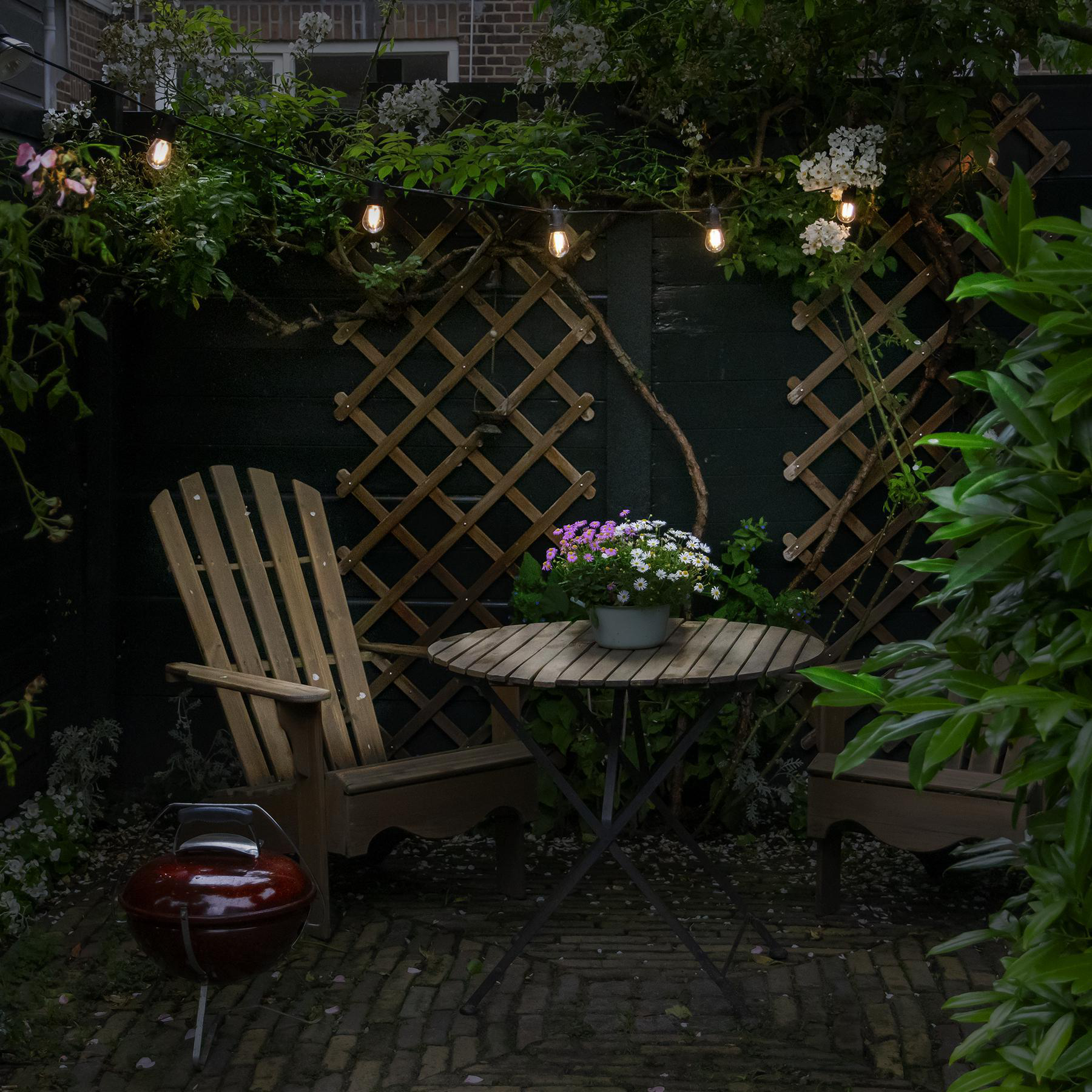 Hinterhofszene mit Holzmöbeln, Lichterketten, Blumen und einem Grill.