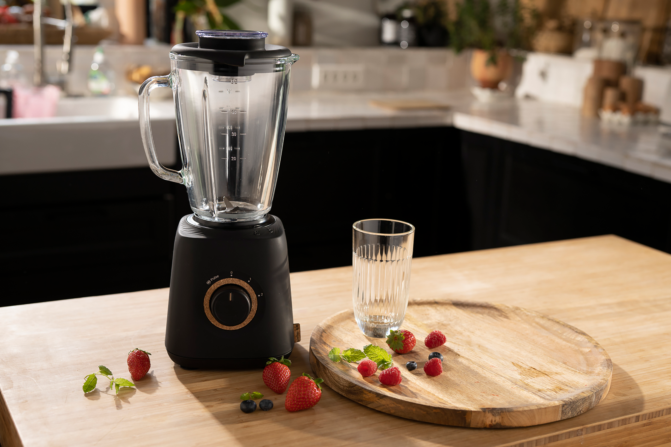 Black blender with a wooden tray, glass, and fruit. Kitchen setting, natural light.