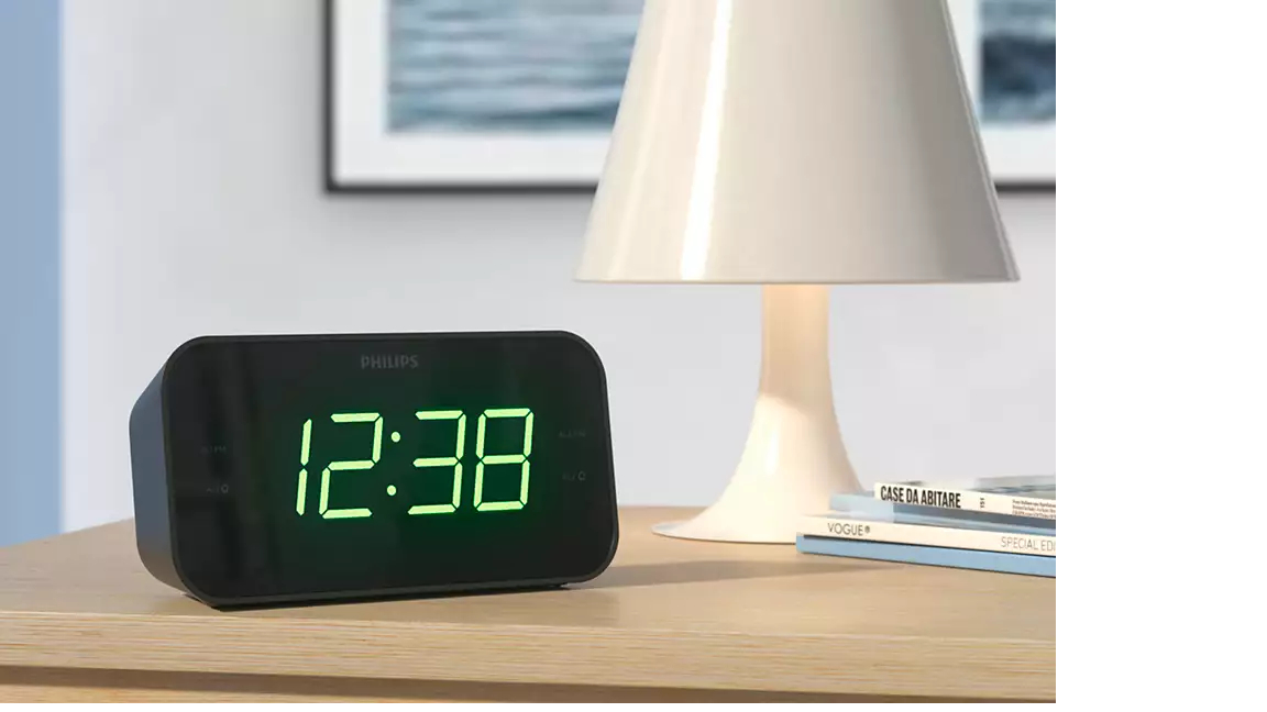 Schwarzer Philips Digitalwecker mit der Anzeige 12:38 auf einem Nachttisch neben einer Lampe.