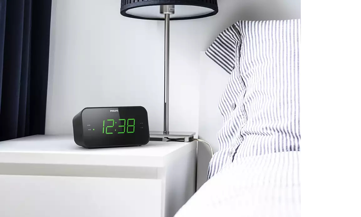 Ein schwarzer digitaler Wecker auf einem weißen Nachttisch neben einem Bett.