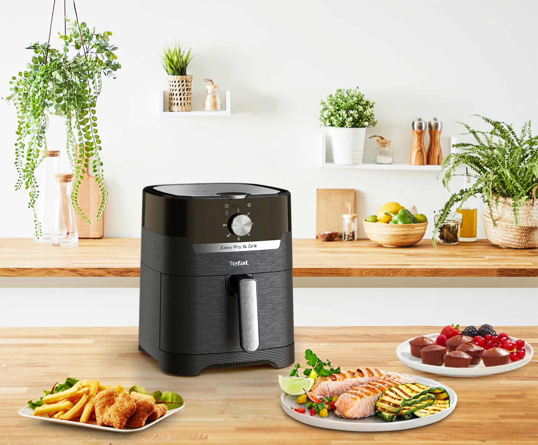 Zwarte airfryer op houten aanrecht met eten. Groenten, friet en muffins aanwezig.